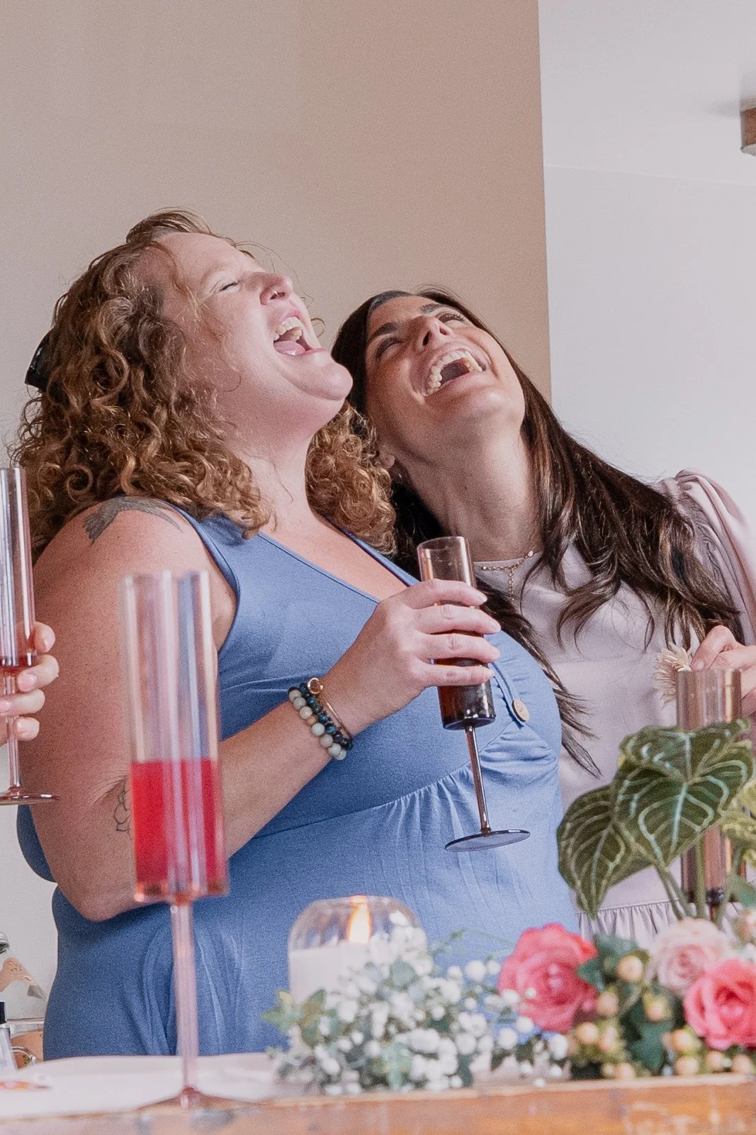 Two women are at a celebration, laughing and holding drinks. The table is decorated with flowers and candles.