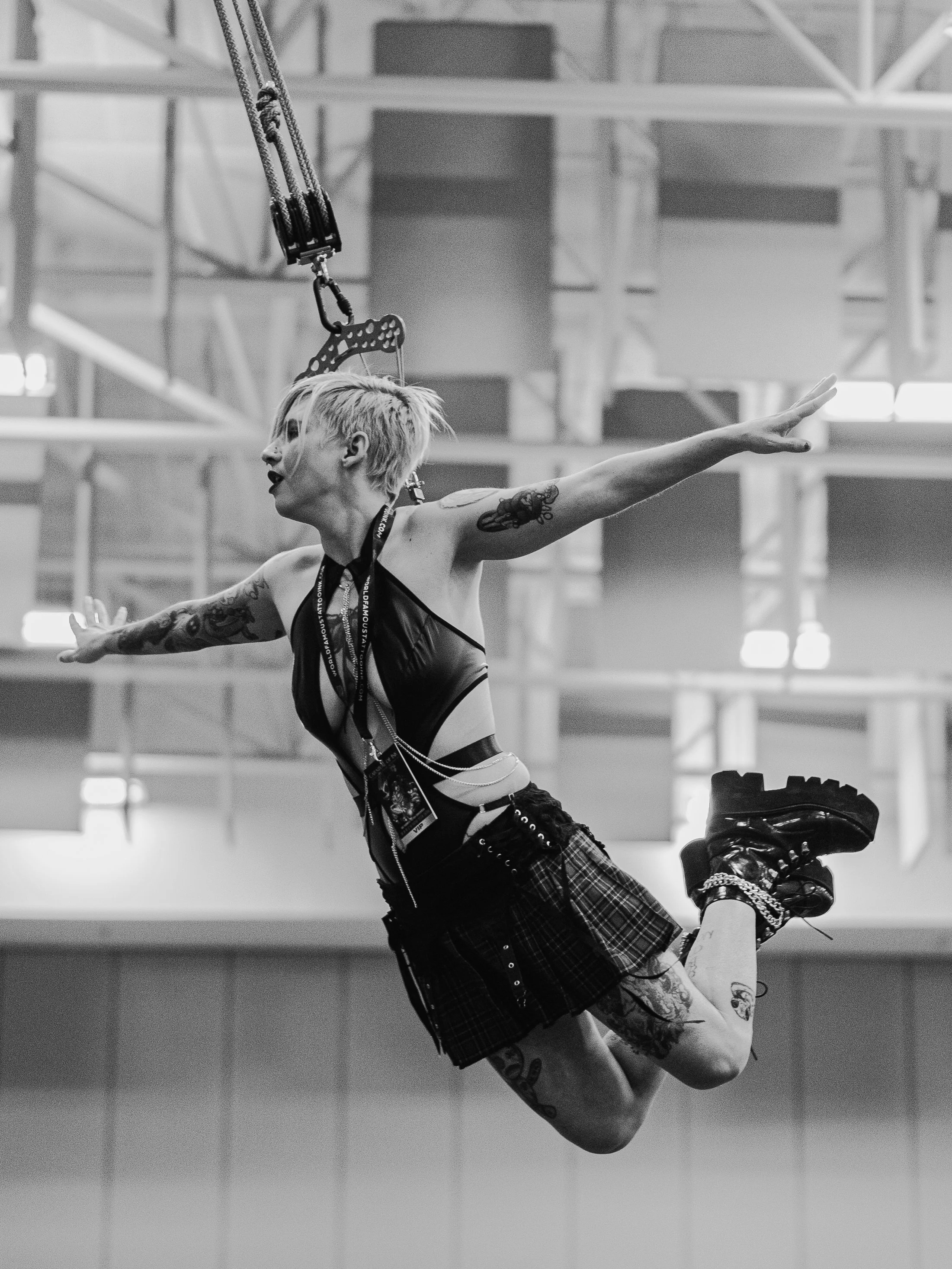 A woman with short, platinum blonde hair and tattoos on her arms, dressed in dark clothing and plaid shorts, performing a split upside down on roller skates in an indoor space with a high ceiling and metal structures.