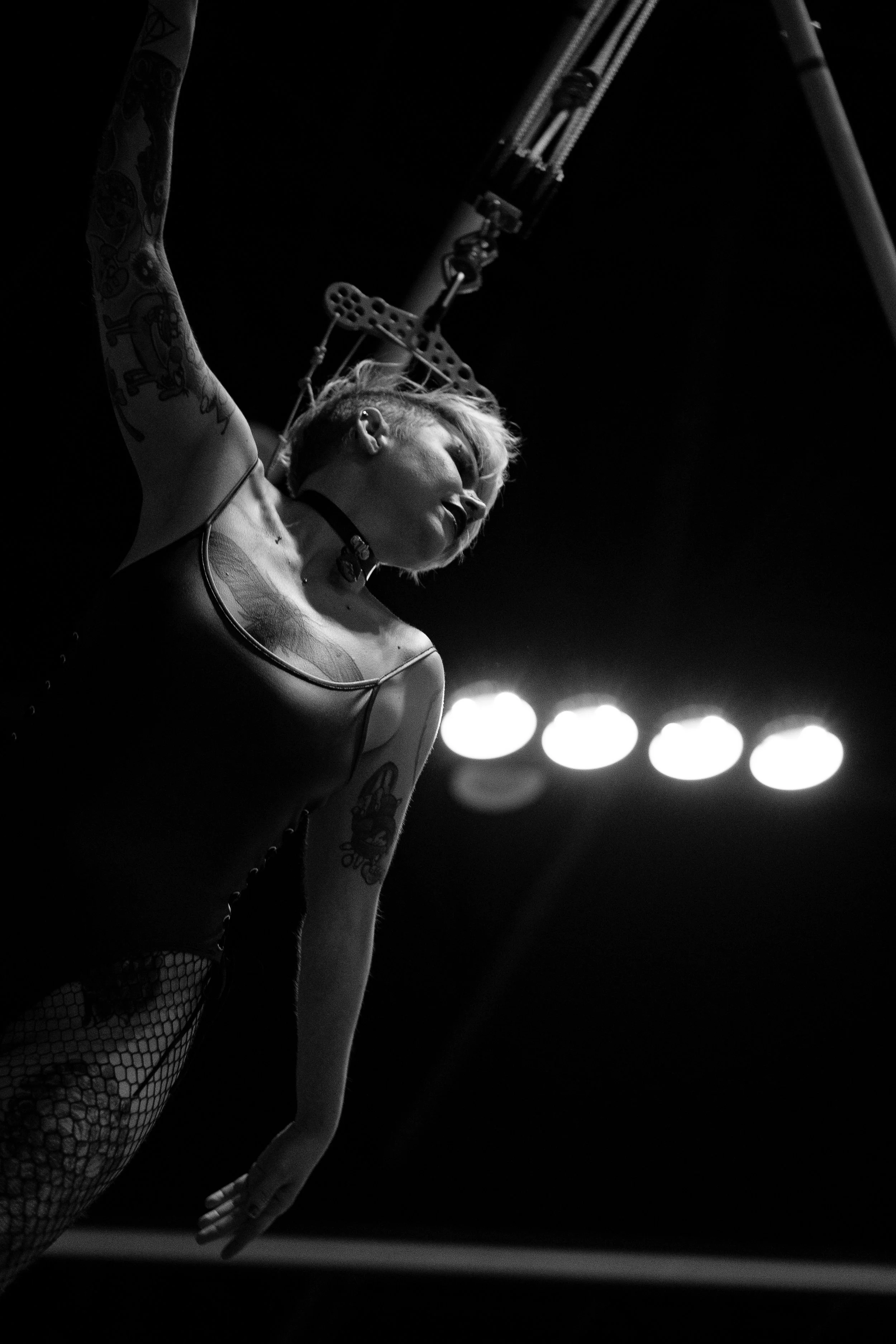 A black and white photo of a woman with tattoos, wearing fishnet stockings and a choker, holding a pole or stick, with stage lights blurred in the background.