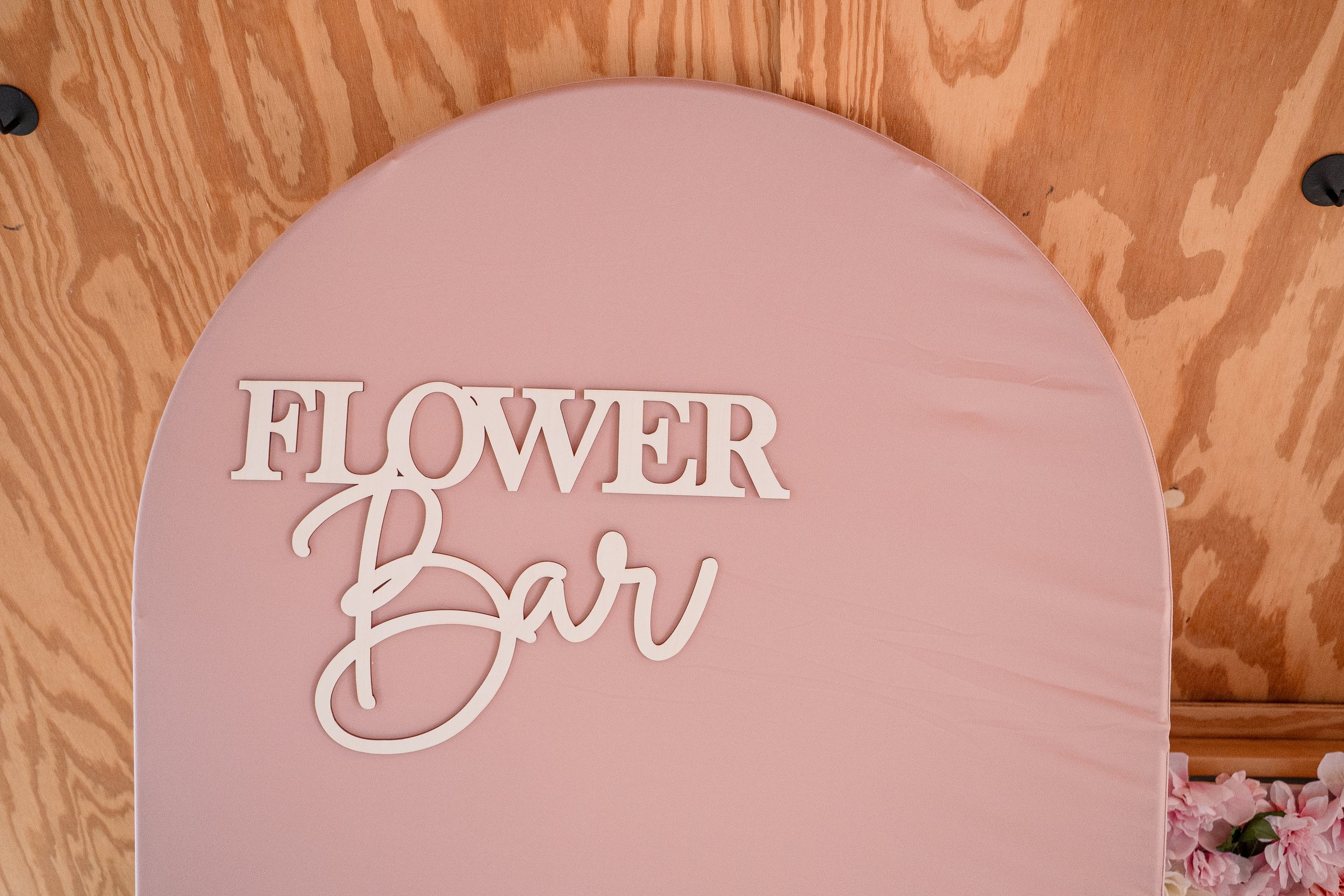 A pink circular sign with the words 'Flower Bar' in decorative white text, placed on a wooden surface with some pink flowers visible at the bottom right corner.