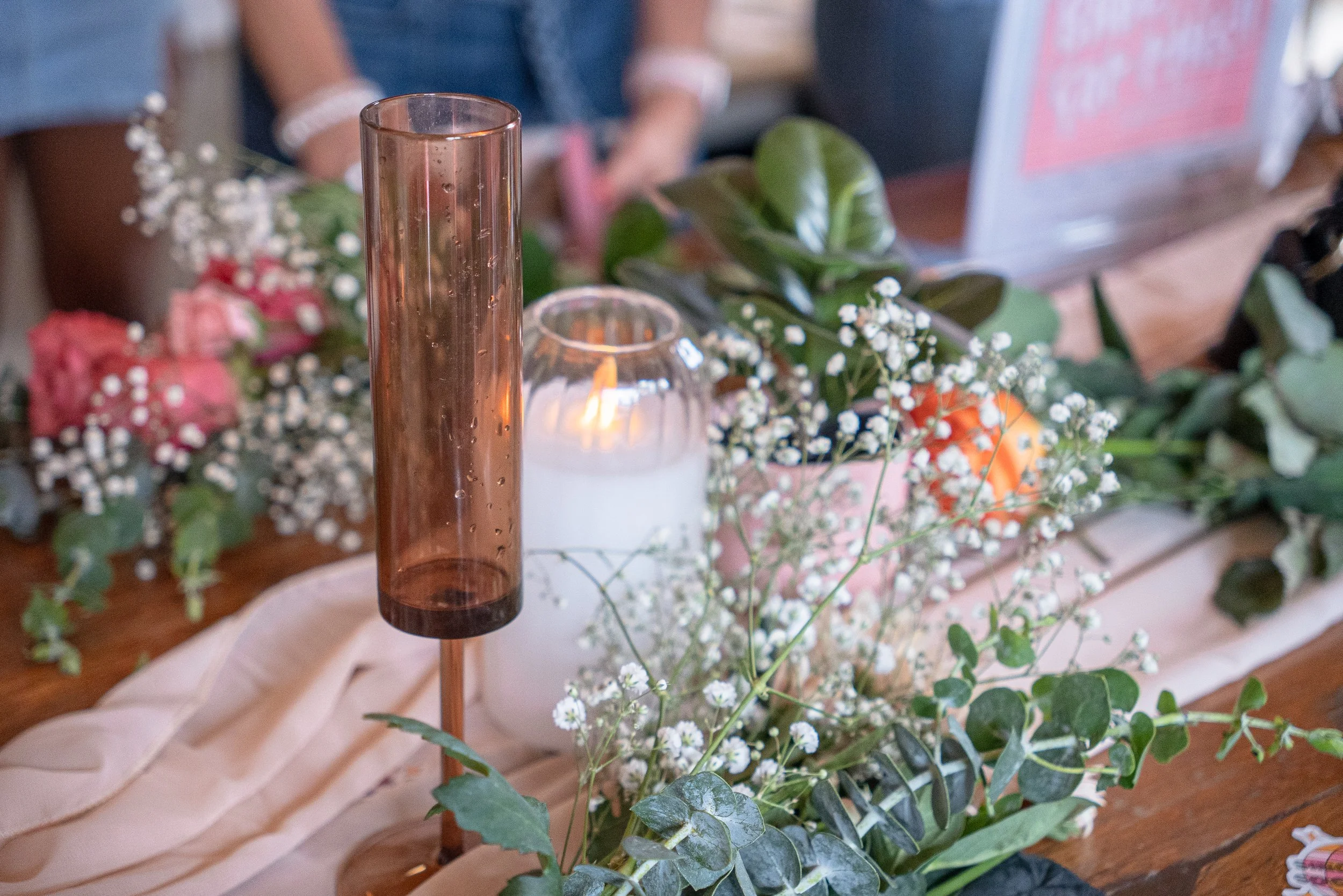 A candle in a tall, cylindrical, brown glass holder surrounded by white baby's breath flowers and green leaves, with a blurred background including a person and a computer.