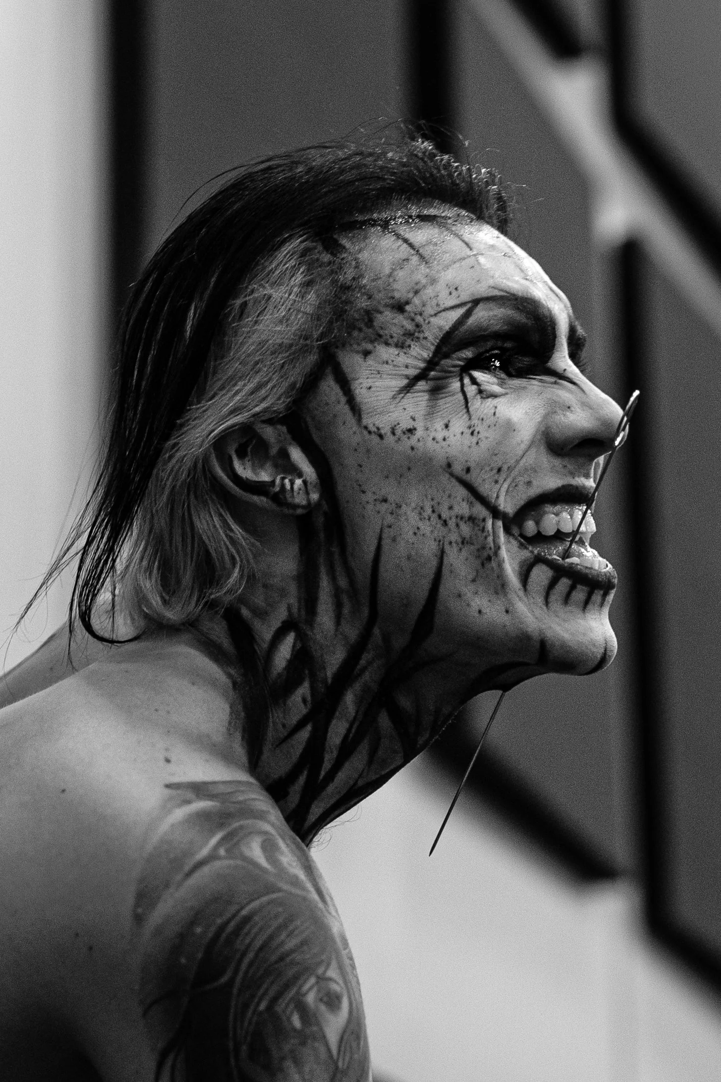 A woman with face paint, earrings, and a tattoo on her arm, smiling and looking to the side in a black and white photo.