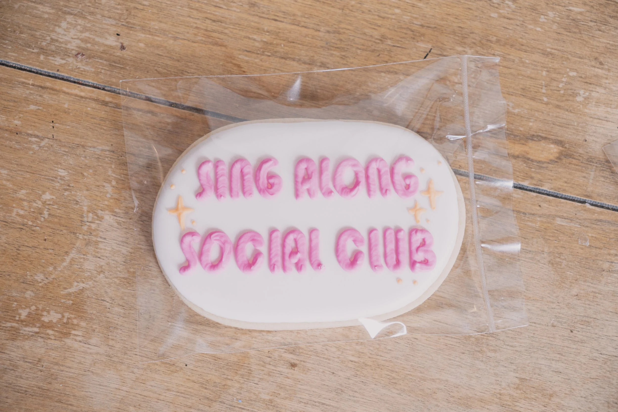 A decorated cookie with pink icing and the words "BIG ALOHA SOCIAL CLUB" written on it, wrapped in clear plastic, resting on a wooden surface.