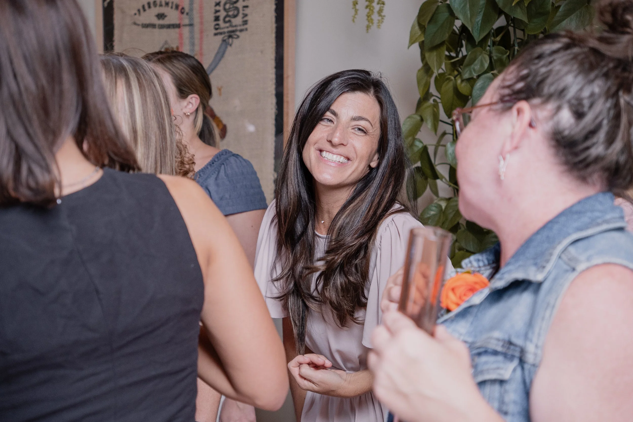 A group of women gathered in a social setting, smiling and engaging in conversation, with one woman in the center smiling warmly.