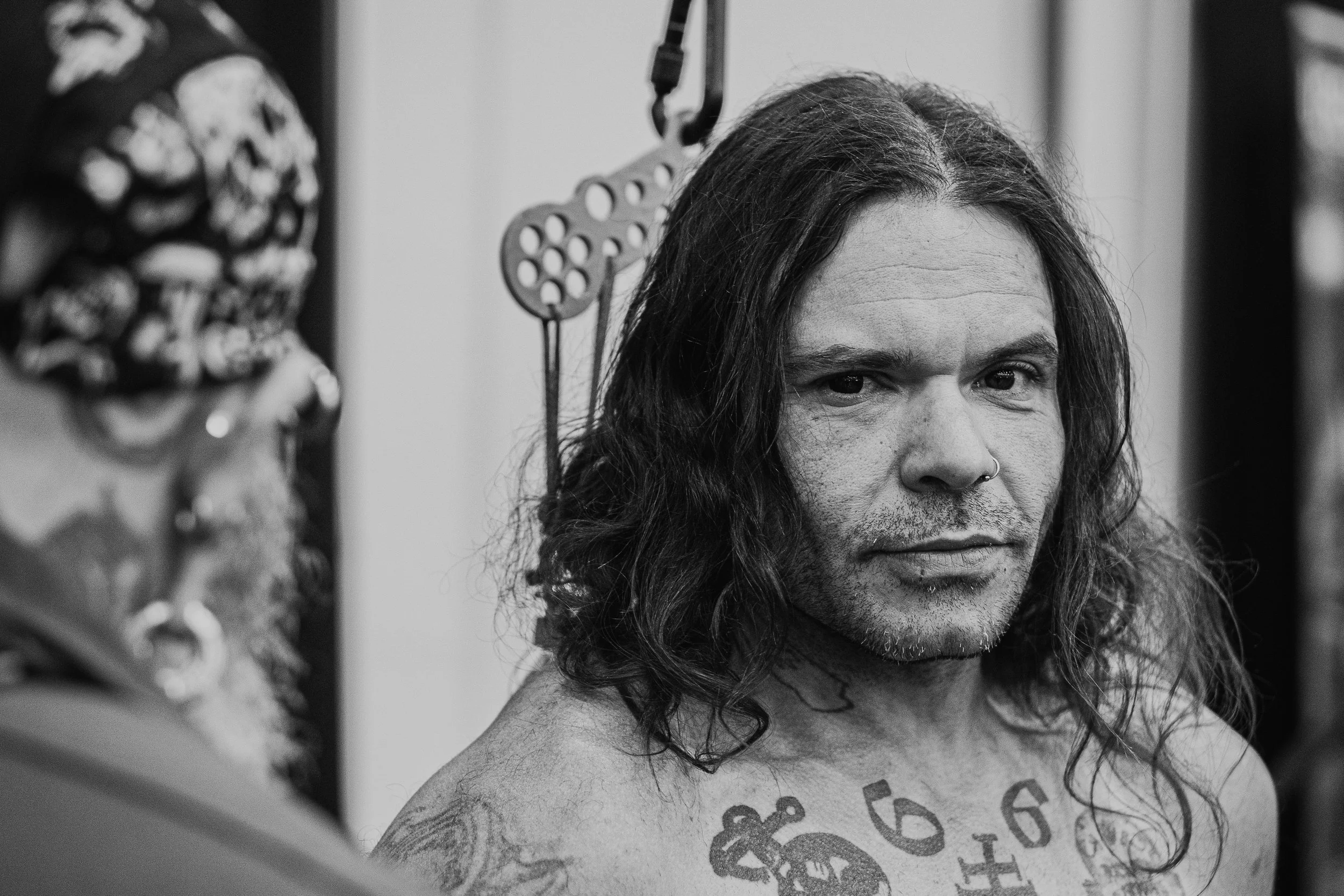 A person with long, wavy hair and a nose ring looking at a camera, displaying tattoos on their chest and shoulder, with a blurred background that includes hanging tools and artwork.