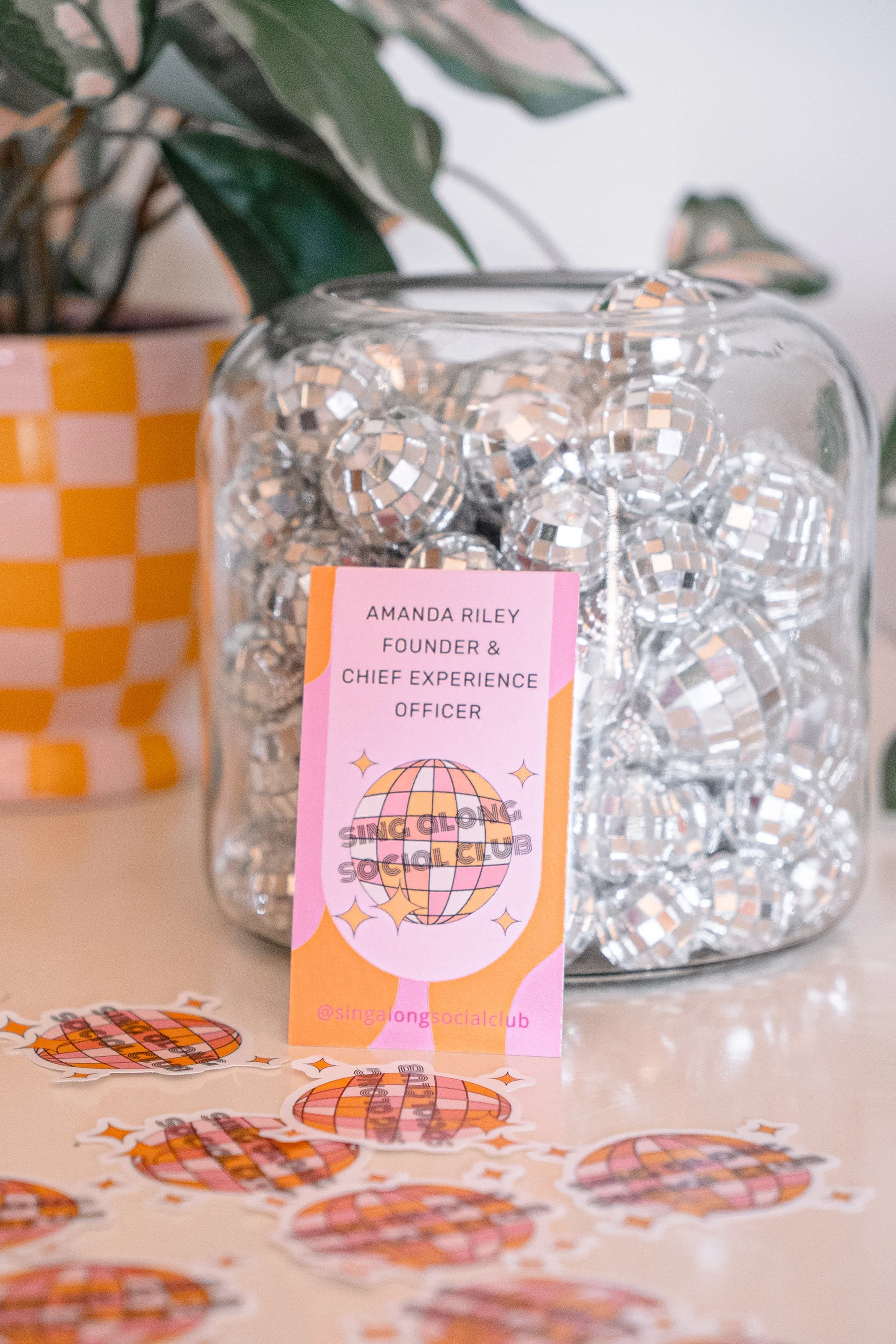 A clear glass jar filled with small silver disco ball decorations. There is a pink and orange tag attached to the jar with the text "Amanda Riley Founder & Chief Experience Officer" and a logo for the Sing Along Social Club featuring a disco ball and