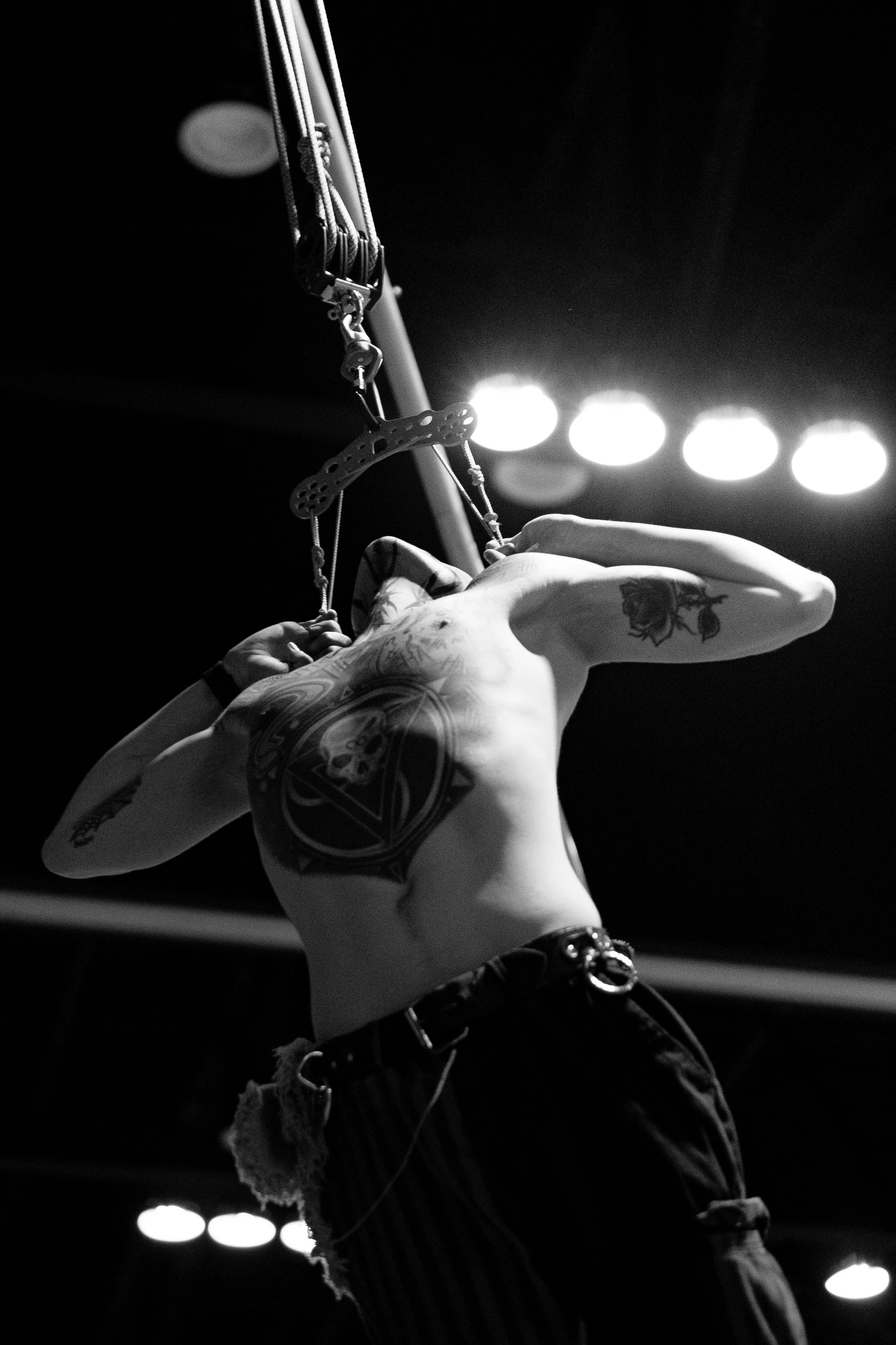 A person's upper body with tattoos on their arm, black wristwatch, and earrings, holding a guitar pick, playing a guitar on stage with bright lights in the background.