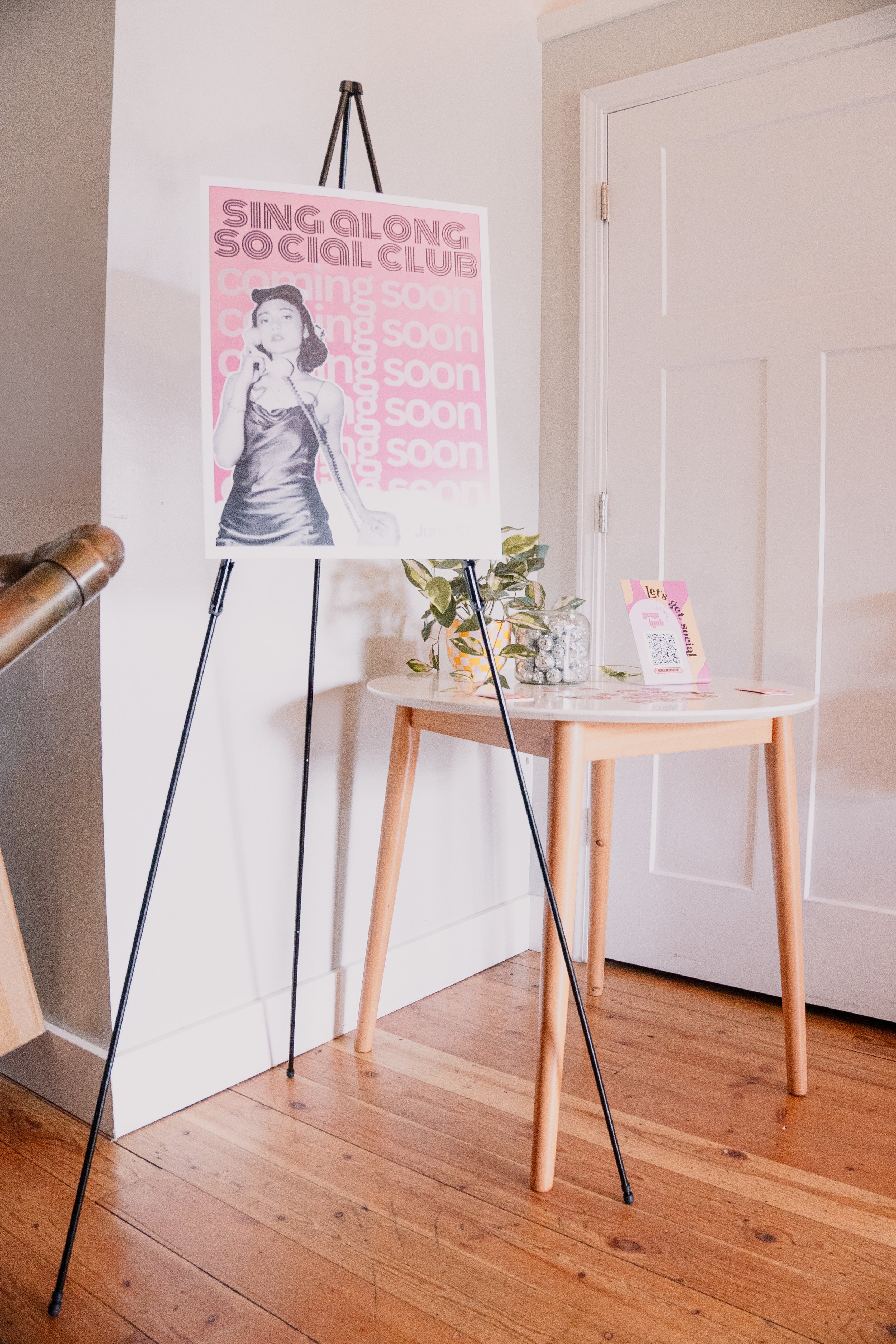 A pink and black poster on an easel features a black-and-white photo of a woman in a dress with dark hair styled in two buns, speaking on a vintage telephone. The poster displays the text 'SING ALONG SOCIAL CLUB' at the top and 'coming soon' repeated
