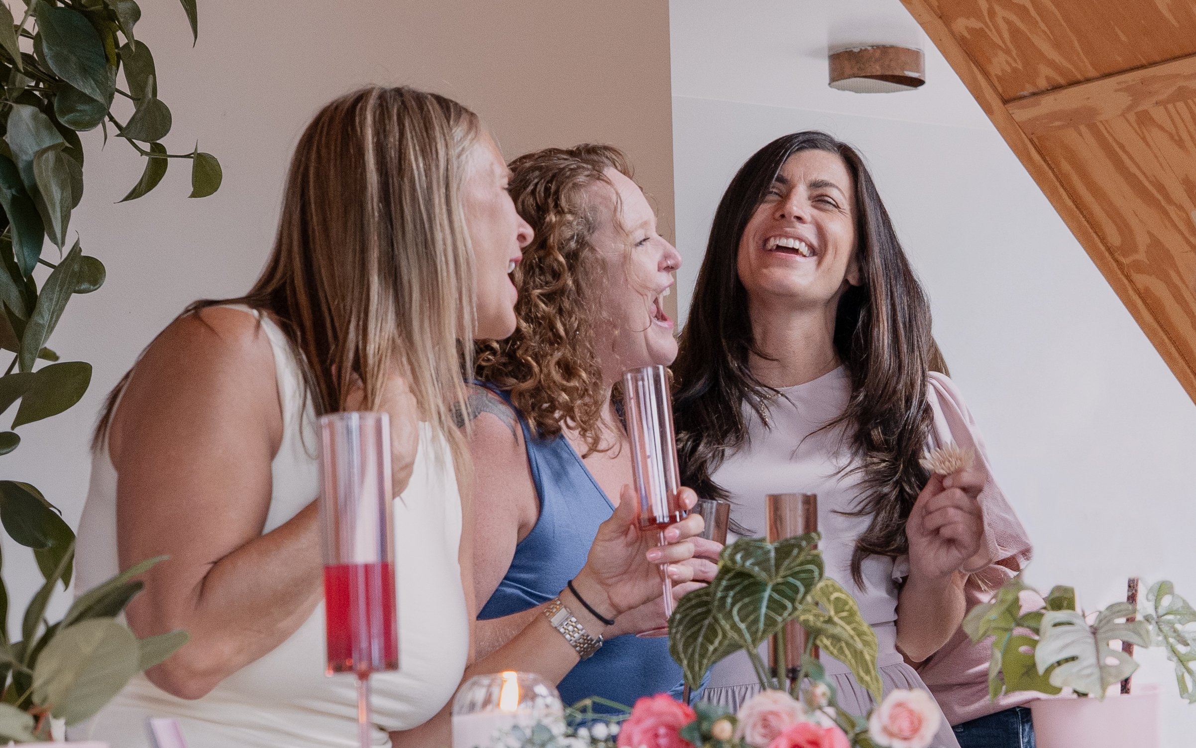 Four women standing together at a celebration, smiling, holding glasses, surrounded by plants and a decorated table with flowers and candles.