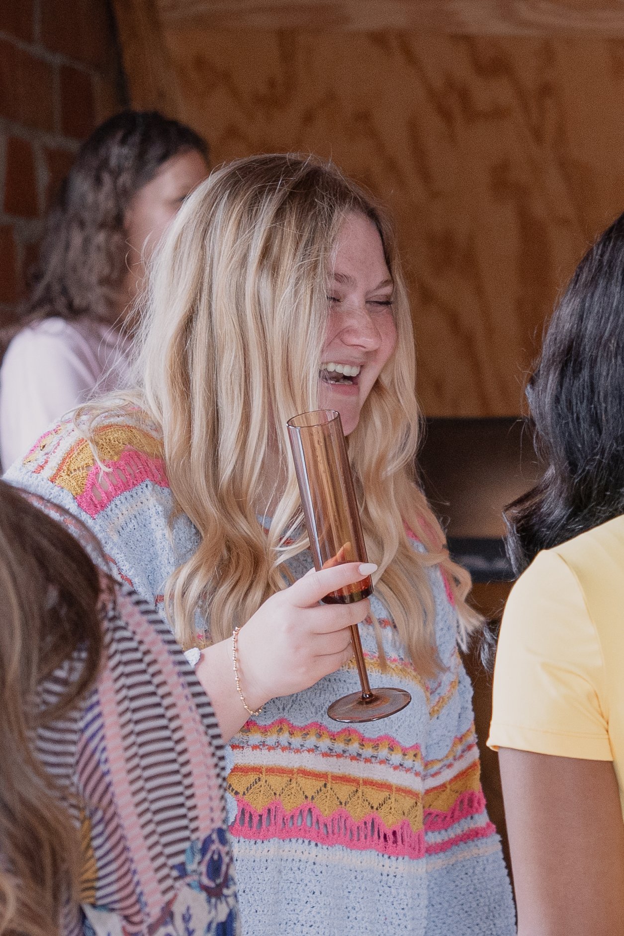 A woman with long blonde hair is holding a tall, empty rose-colored champagne flute and smiling at a social gathering. She is wearing a colorful, patterned sweater and has a bracelet on her wrist. Other people are partially visible around her, and th