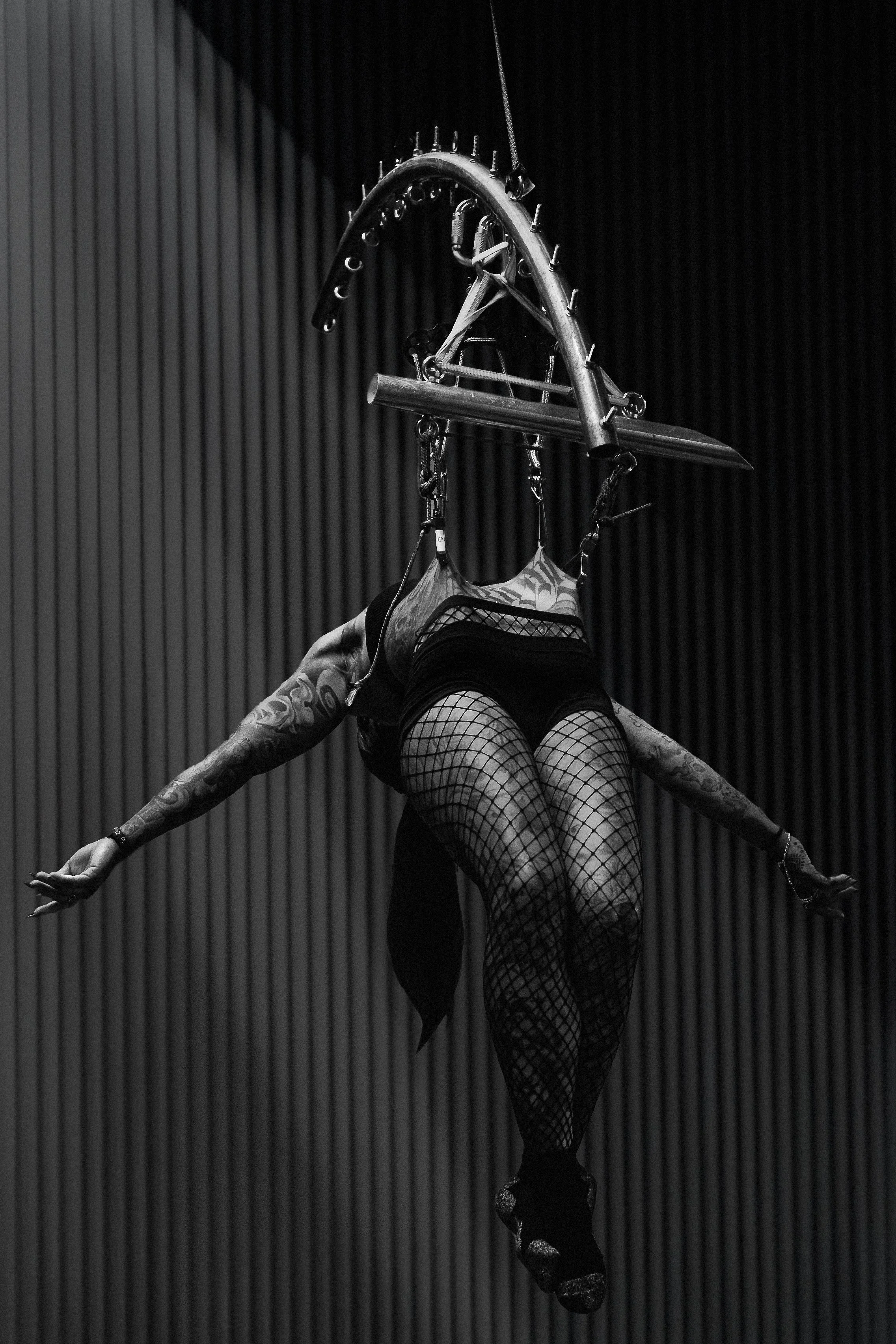 A performer in fishnet stockings and fishnet gloves is on stage, hanging upside down, holding a sword with one hand. The sword's handle is secure on a metal overhead structure with various attachments. The background consists of vertical black panels