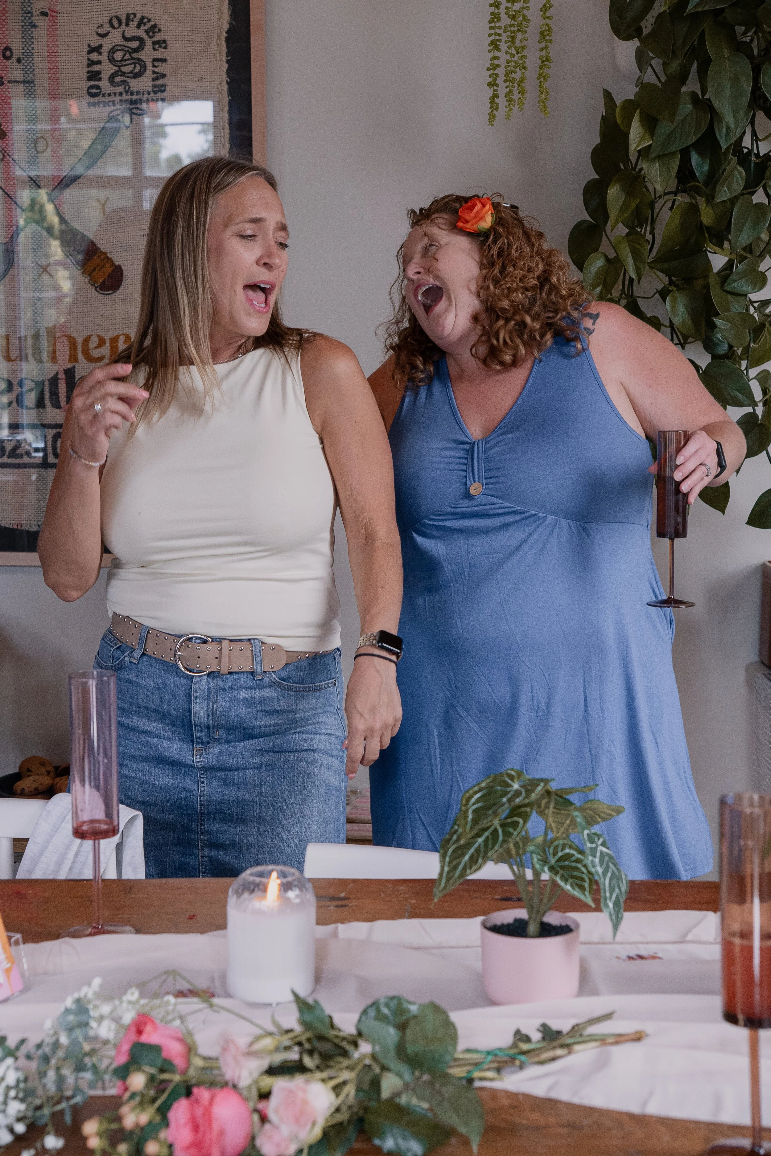 Two women singing together at a celebration or party, standing behind a decorated table with candles, flowers, and greenery.