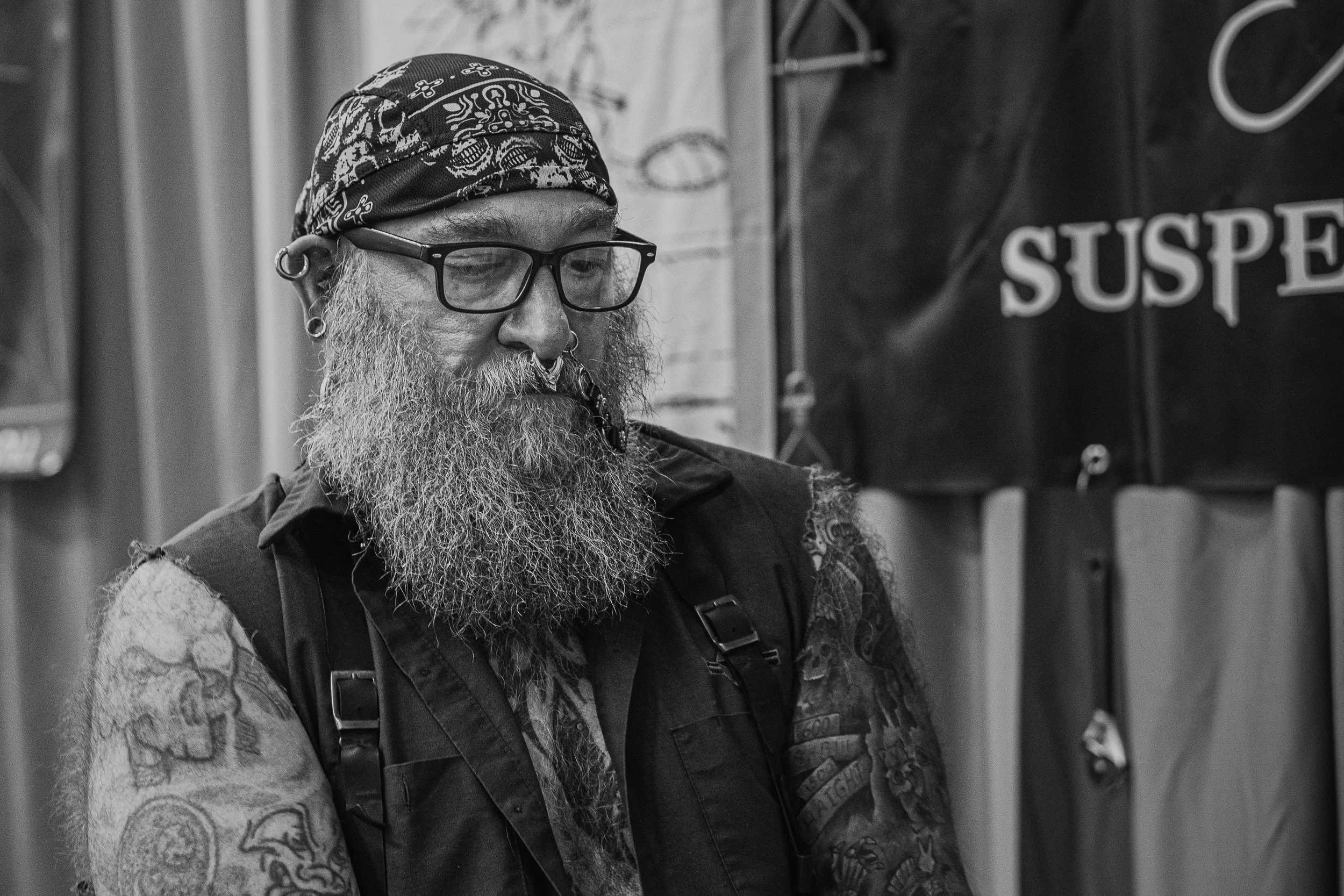 A bearded man wearing glasses, a bandana on his head, and tattoos on his arms is seen in a black-and-white photo. He is wearing a vest with shoulder straps and appears to be looking down or reading.