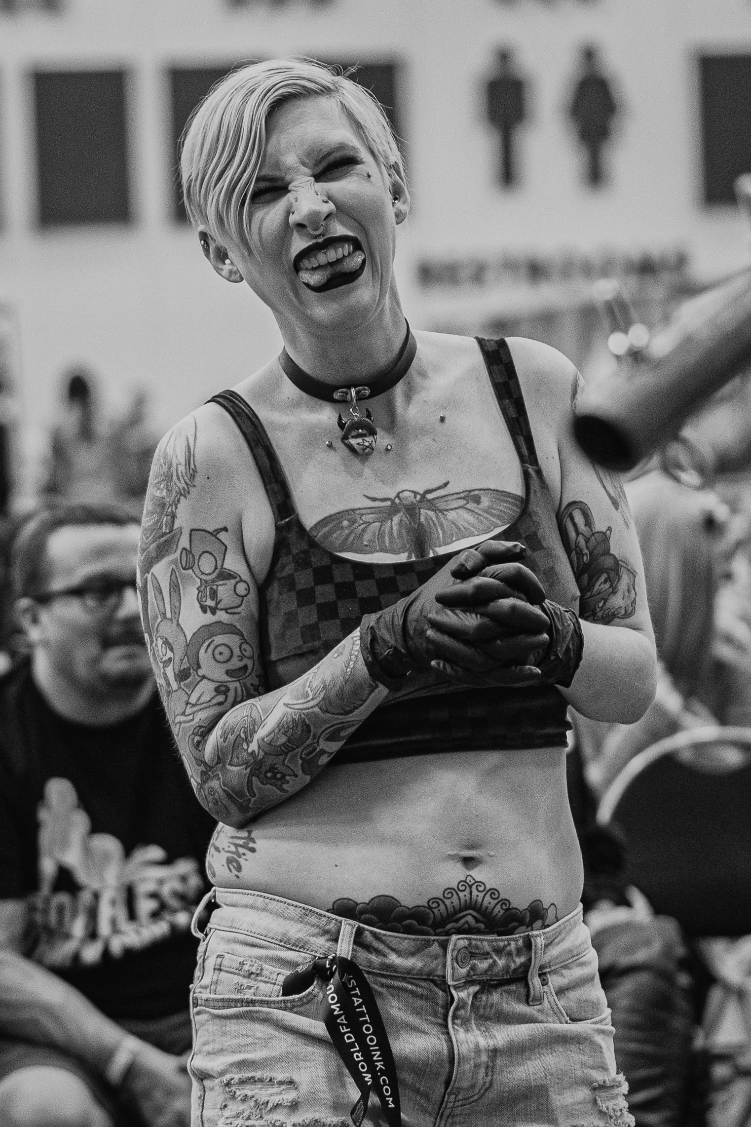 A woman with short platinum blonde hair and tattoos on her arms and torso, sticking her tongue out and making a fierce expression. She is wearing a choker with a pendant, a checkered sleeveless top, and ripped denim shorts. The background is blurred 