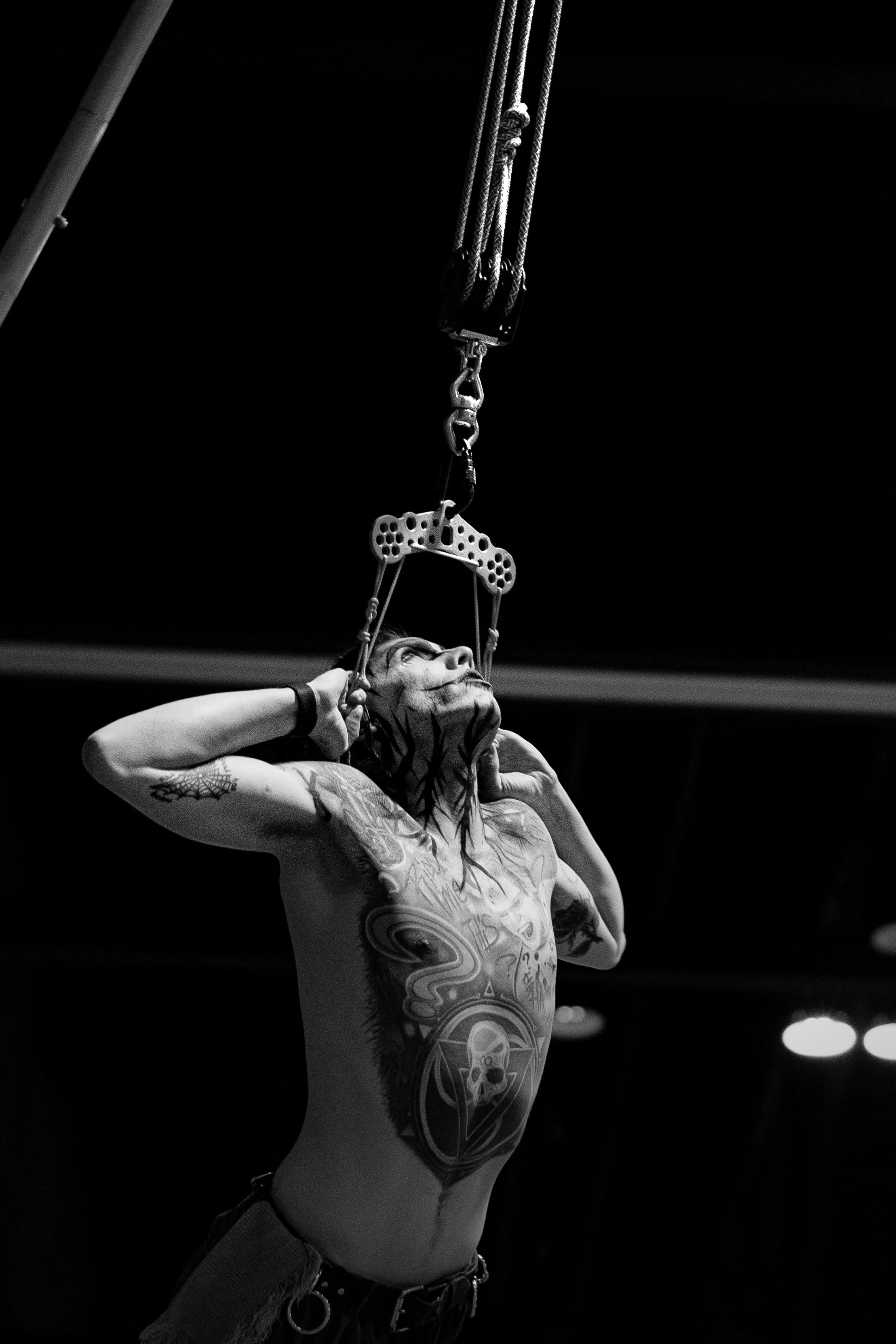 A tattooed man with long hair is holding a hook attached to a pulley system, possibly in a training or performance setting, with a dark background.