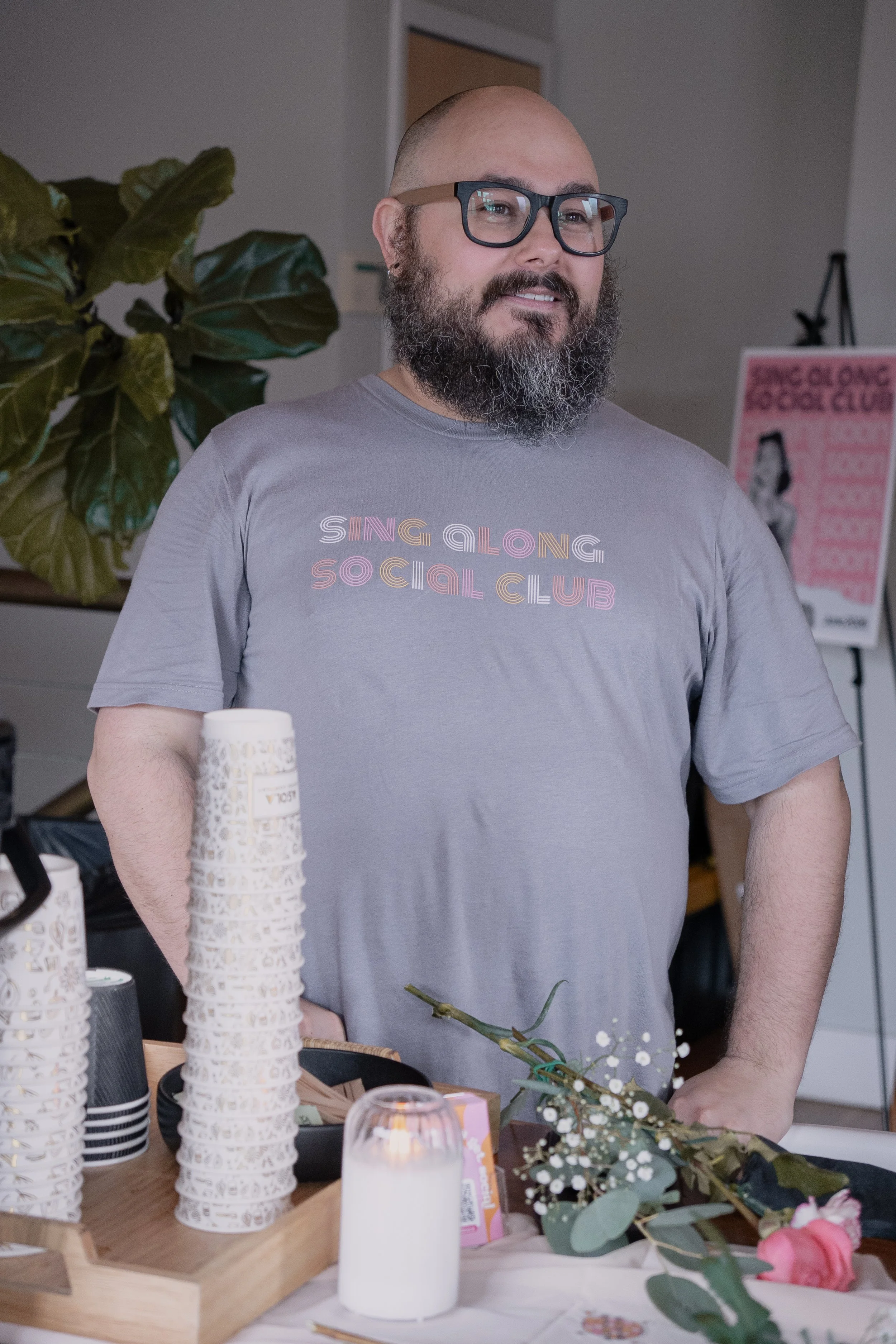 A man with glasses, a beard, and a shaved head standing behind a table with stacked paper cups, flowers, and a candle.