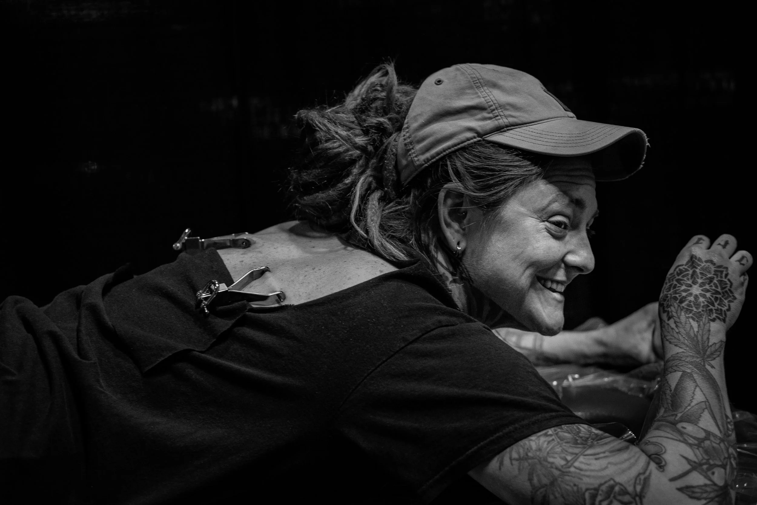 A woman with curly hair wearing a baseball cap and dark shirt smiling and reaching with her tattooed arm.