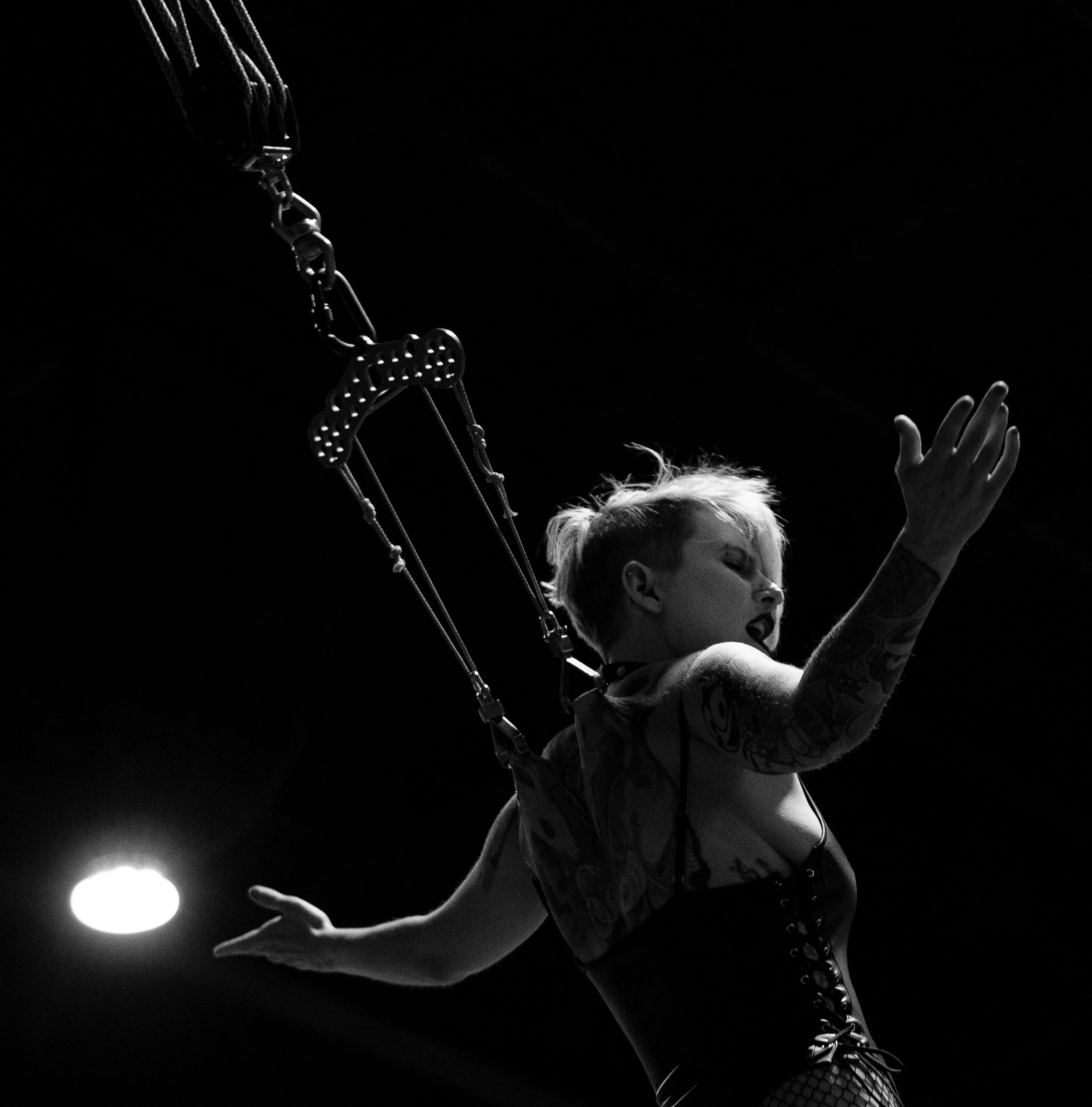 A young woman singing or speaking passionately on stage, illuminated against a dark background. She has blonde hair, tattoos on her arm, and is reaching upward with her hand.