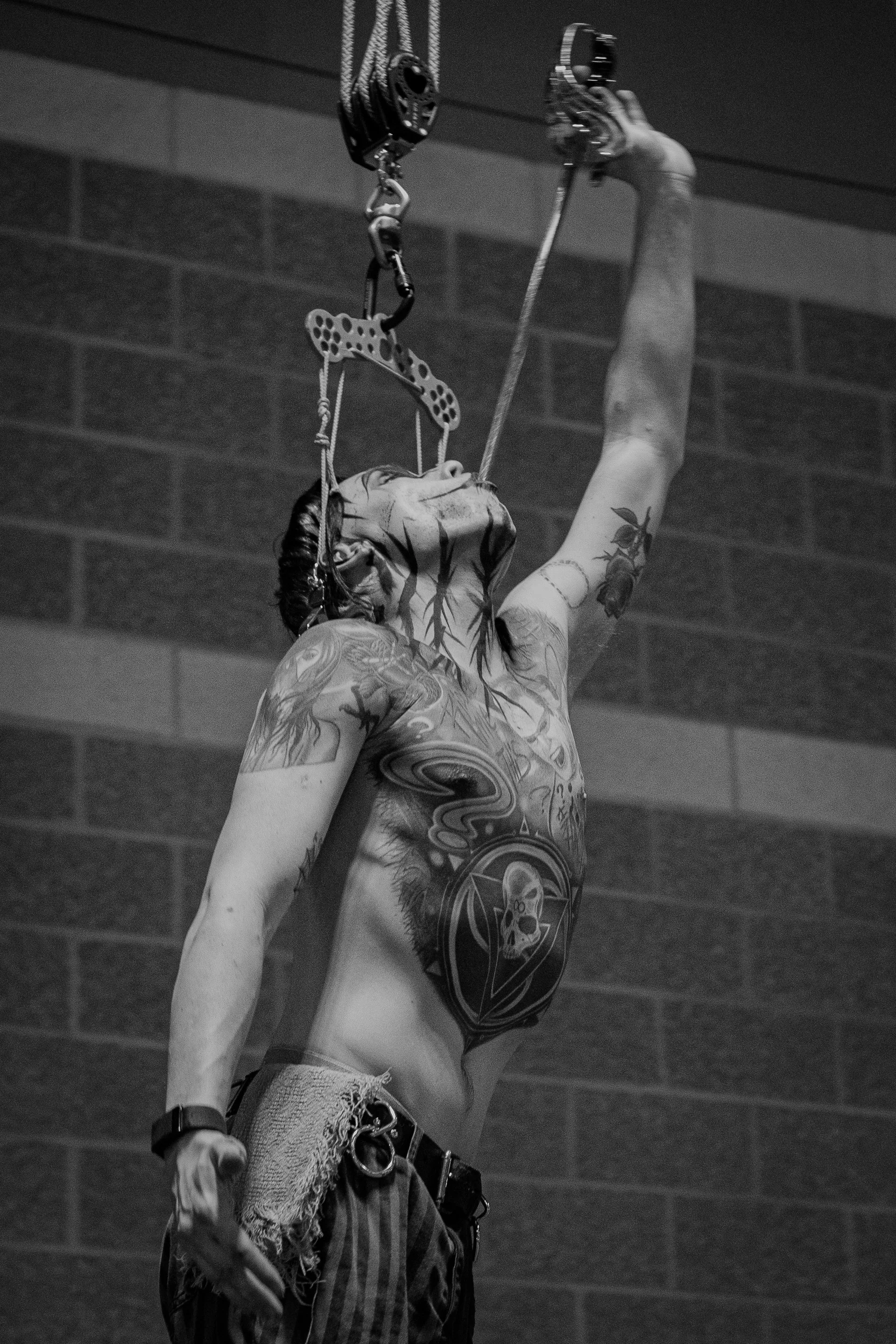 A person with tattoos on their chest and arms, wearing ripped jeans and a wristwatch, is reaching up to adjust climbing hardware attached to a ceiling hook, with a brick wall in the background.