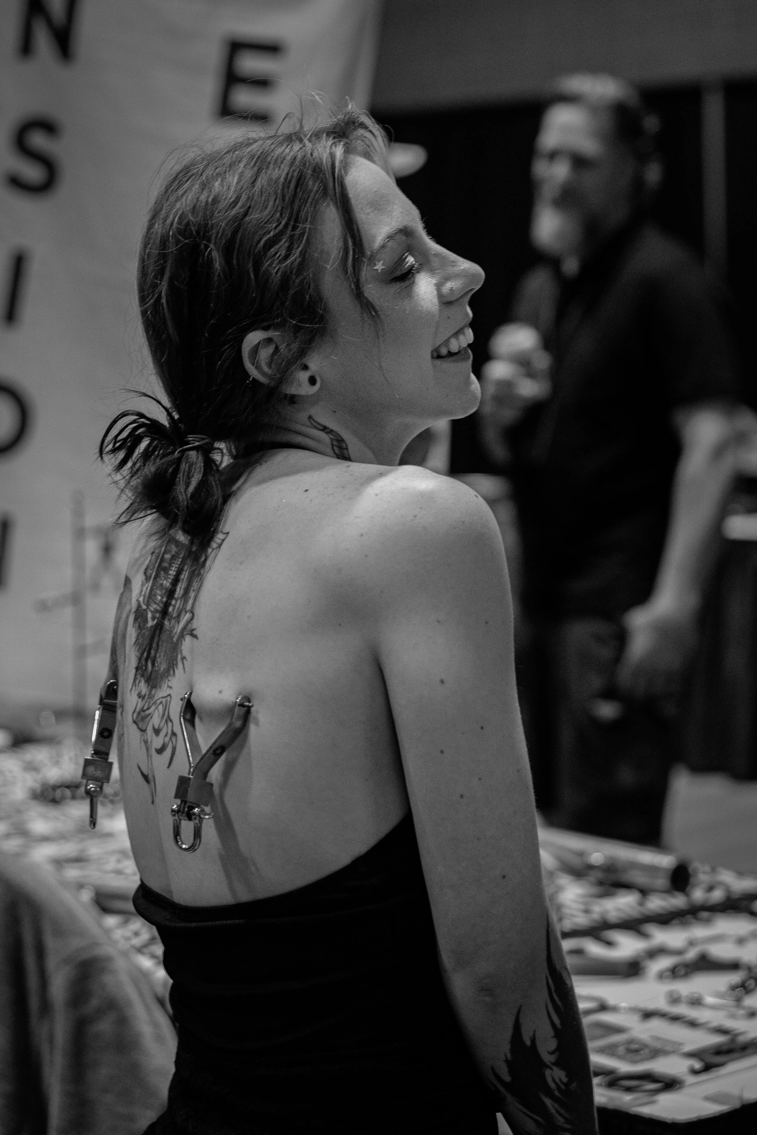 A young woman with tattoos on her back and a smile, sitting at a table with tattoos and art supplies, while an older man in the background watches and talks.