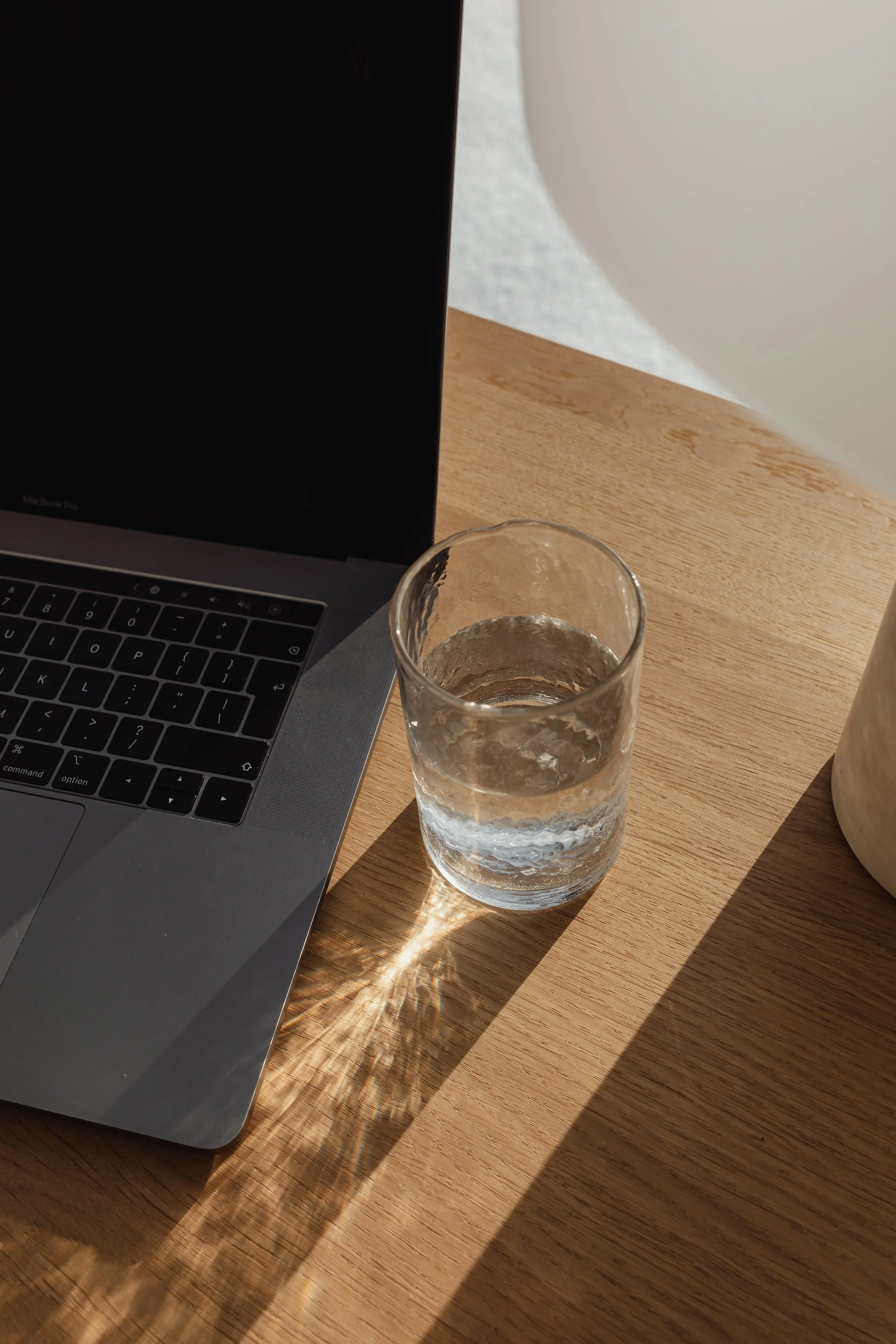 Een open MacBook laptop op een houten tafel, naast een glas water dat door het licht een schaduw en reflectie op de tafel werpt. In de achtergrond een witte muur.
