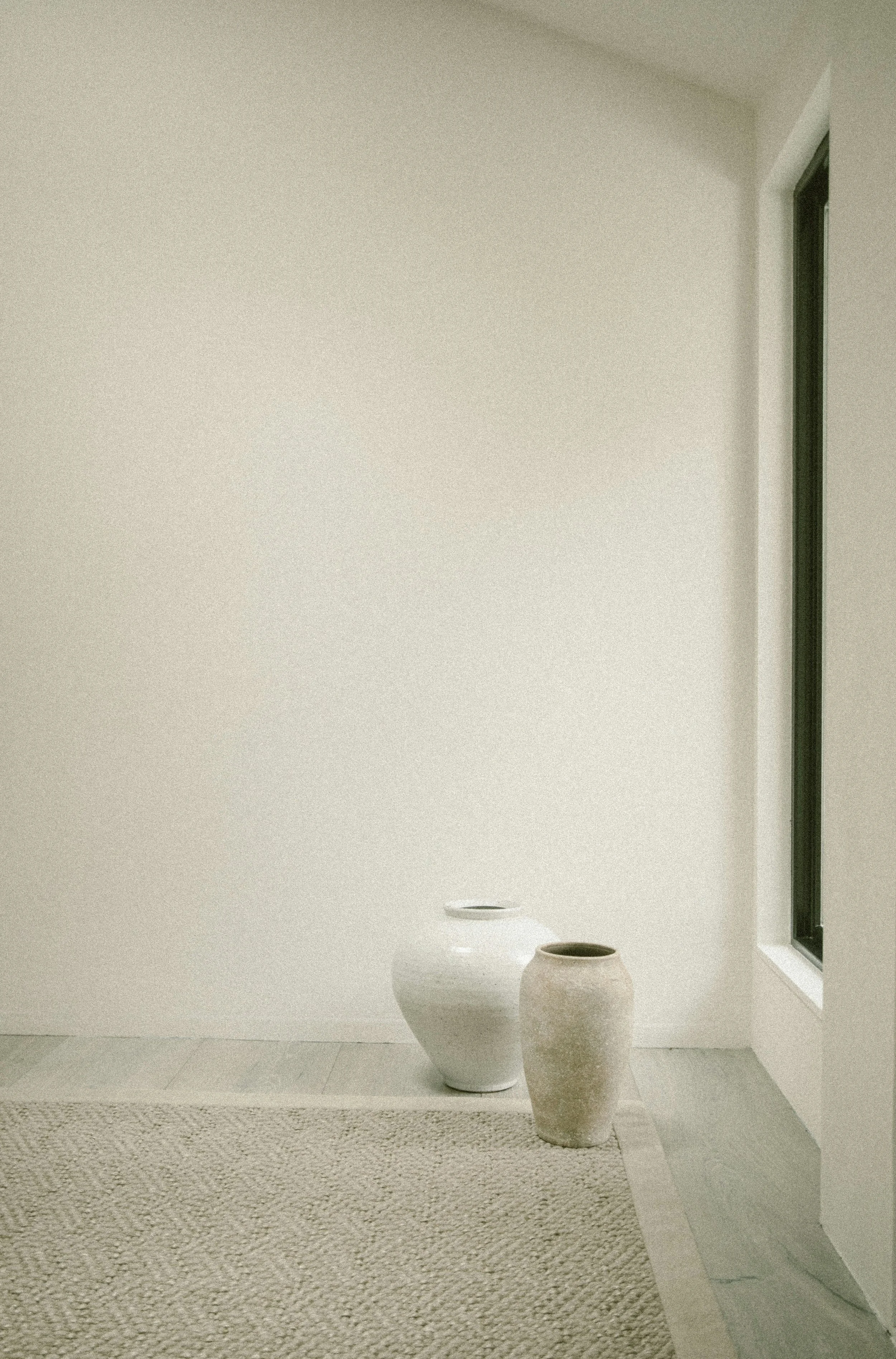 A minimalist interior scene with two large white vases on a tiled floor, near a window with a black frame, against a plain off-white wall.