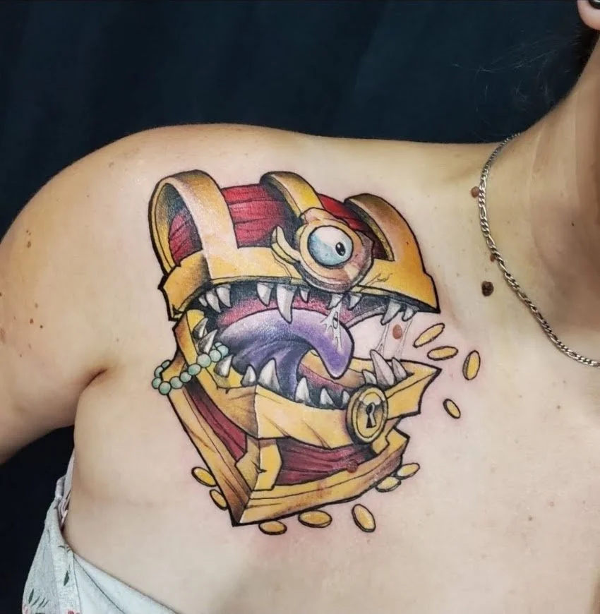 Colorful tattoo of a cartoonish treasure chest with a large open mouth, sharp teeth, a single eye, and gold coins spilling out on the chest of a person.