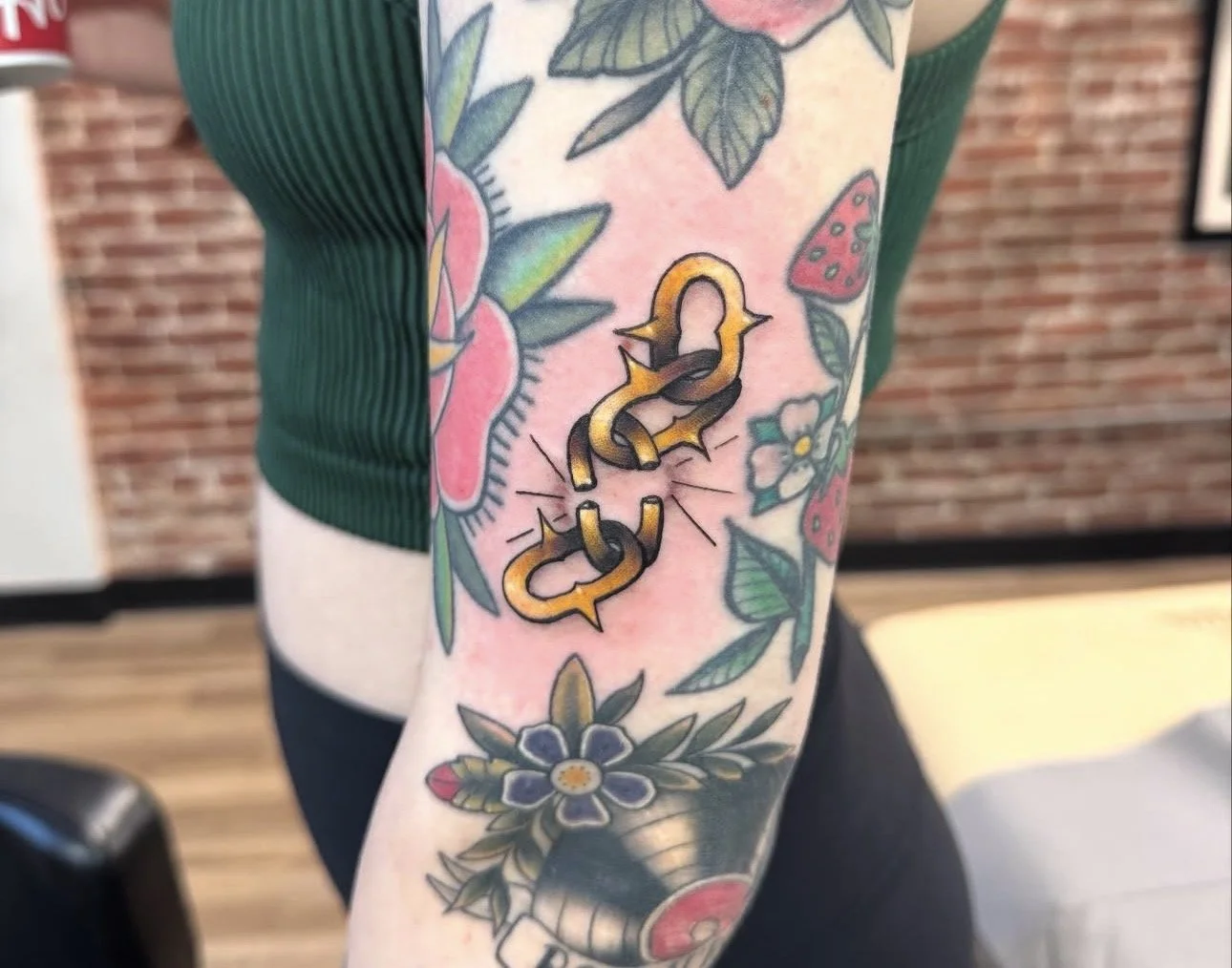 Tattoo on person's arm featuring a chain link design in yellow and black, surrounded by colorful flowers and leaves against a pink background.