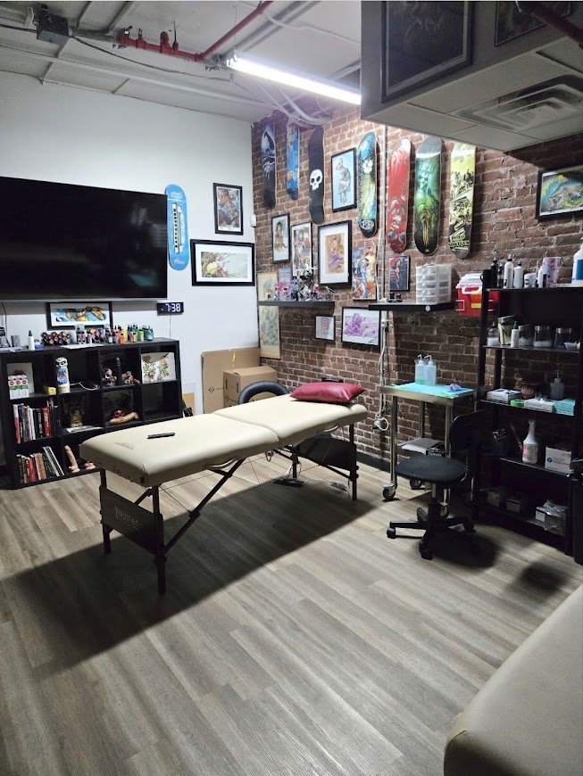 A tattoo studio with a massage or treatment table in the center, decorated with framed artwork, skateboards on the brick wall, and shelves with art supplies and books.