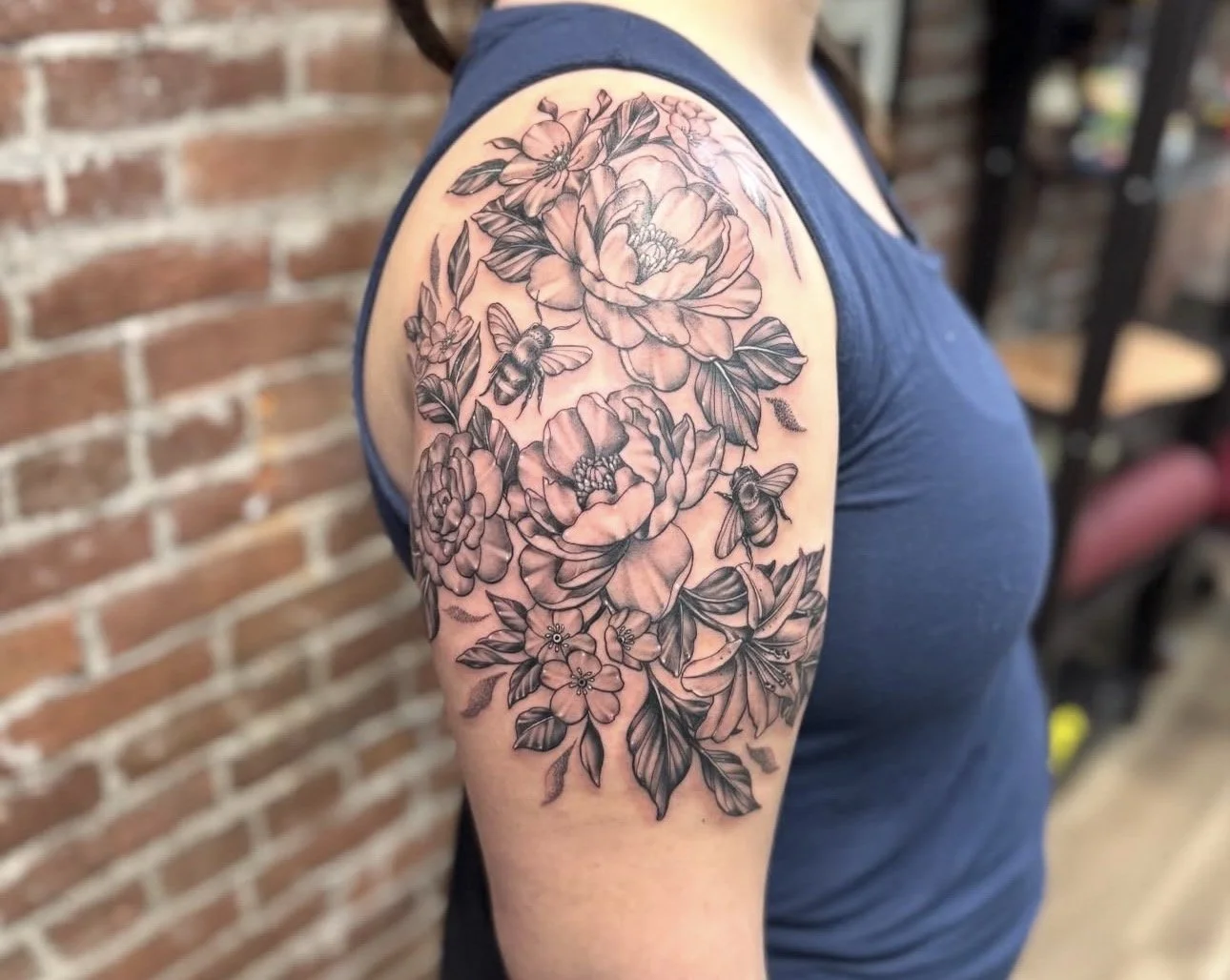 Black and gray floral tattoo on upper arm featuring large flowers, leaves, and bees.