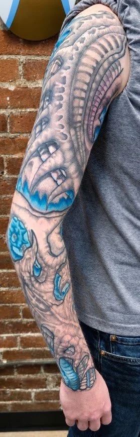 Tattoo sleeve on person's arm featuring a dragon and abstract blue elements against a brick wall. bio mechanical 