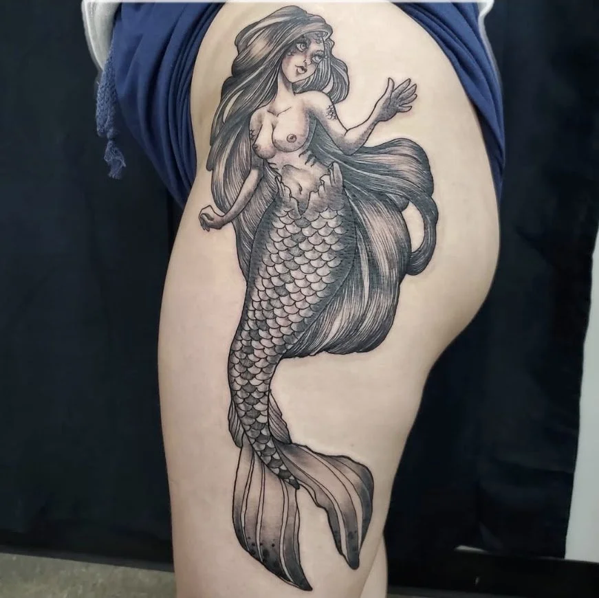 A detailed black and gray tattoo of a mermaid with long flowing hair and a scaled tail on a person's thigh.