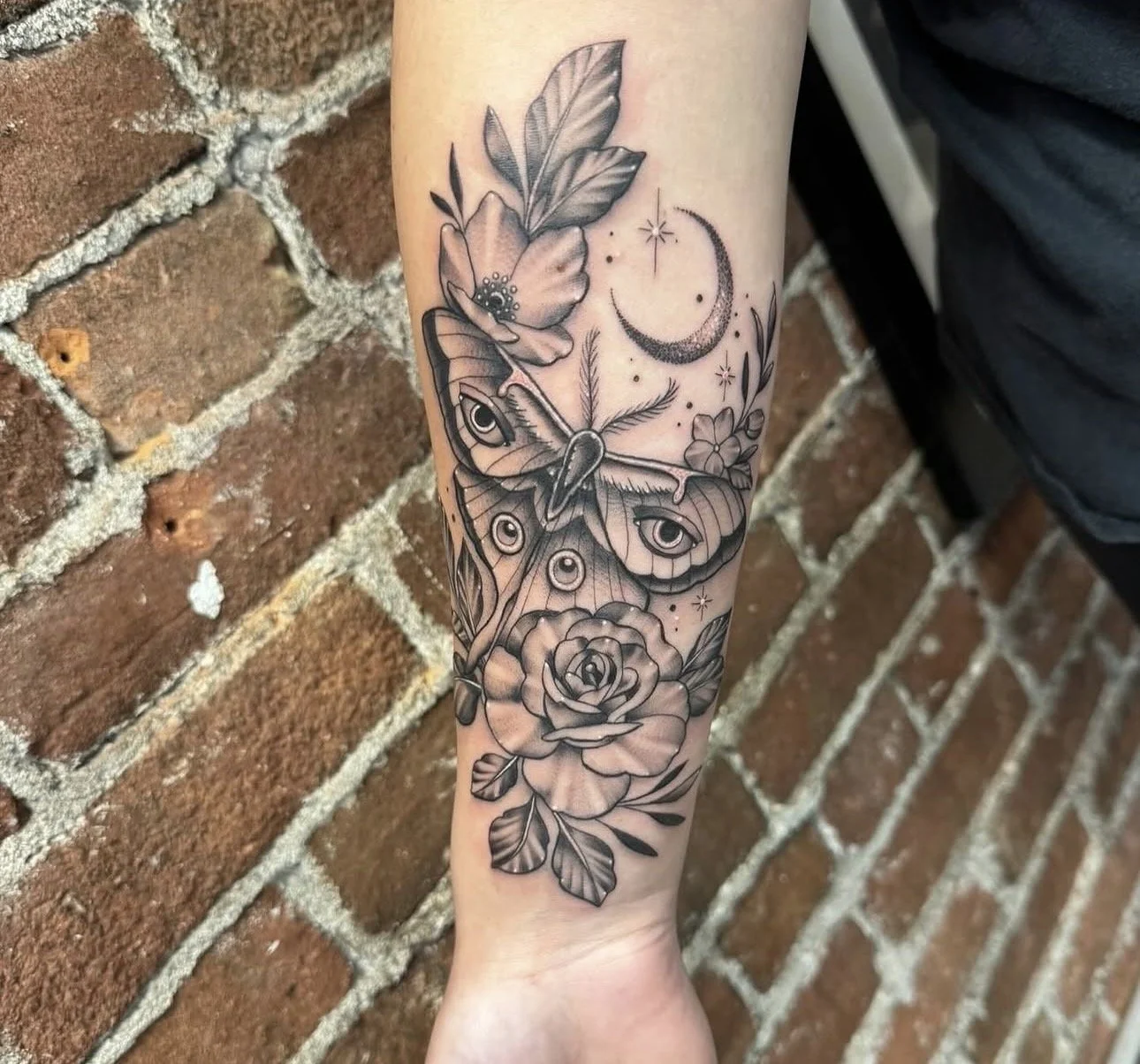 Black and gray floral tattoo featuring a rose, leaves, an eye, a butterfly, and celestial elements like a crescent moon and stars on a person's forearm.