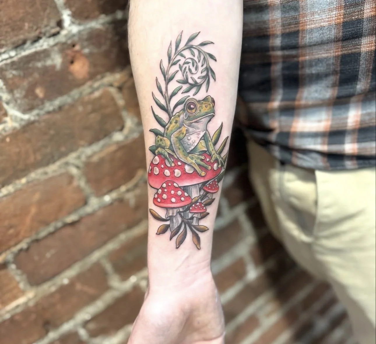 Tattoo of a frog sitting on red and white mushrooms with green foliage and a swirling branch on a person's forearm.