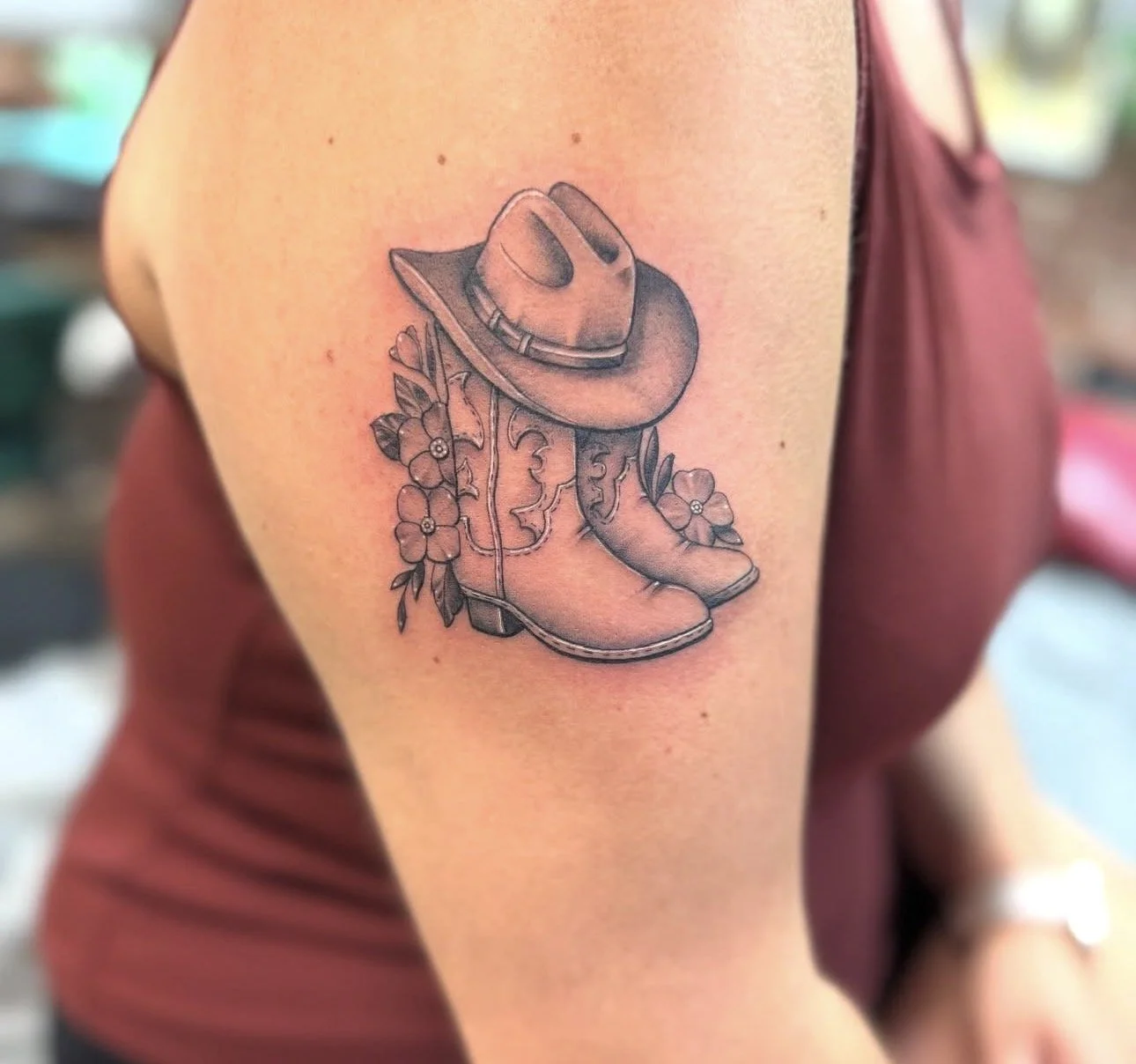 Tattoo of a cowboy hat, cowboy boot, and flowers on a person's upper arm.