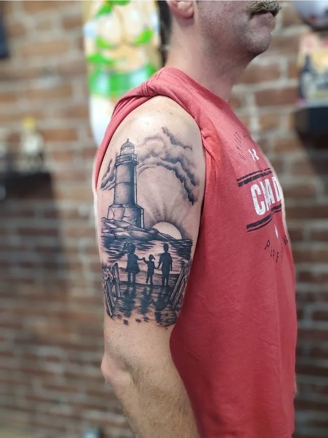 Tattoo of a lighthouse, ocean waves, and three people holding hands at sunset or sunrise on a man's upper arm.