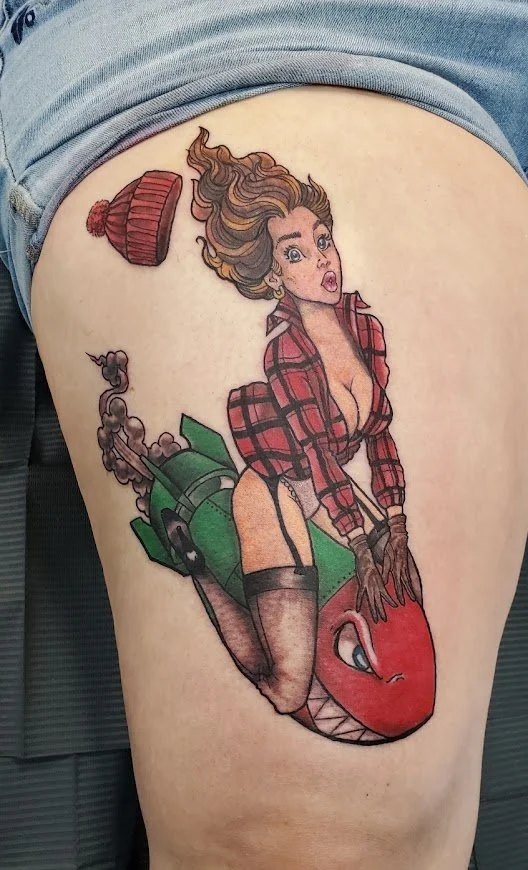 Tattoo of a surprised woman with long hair, wearing a red plaid shirt, sitting on a large anthropomorphic tomato with a face, and a vintage red beanie hat above her head.