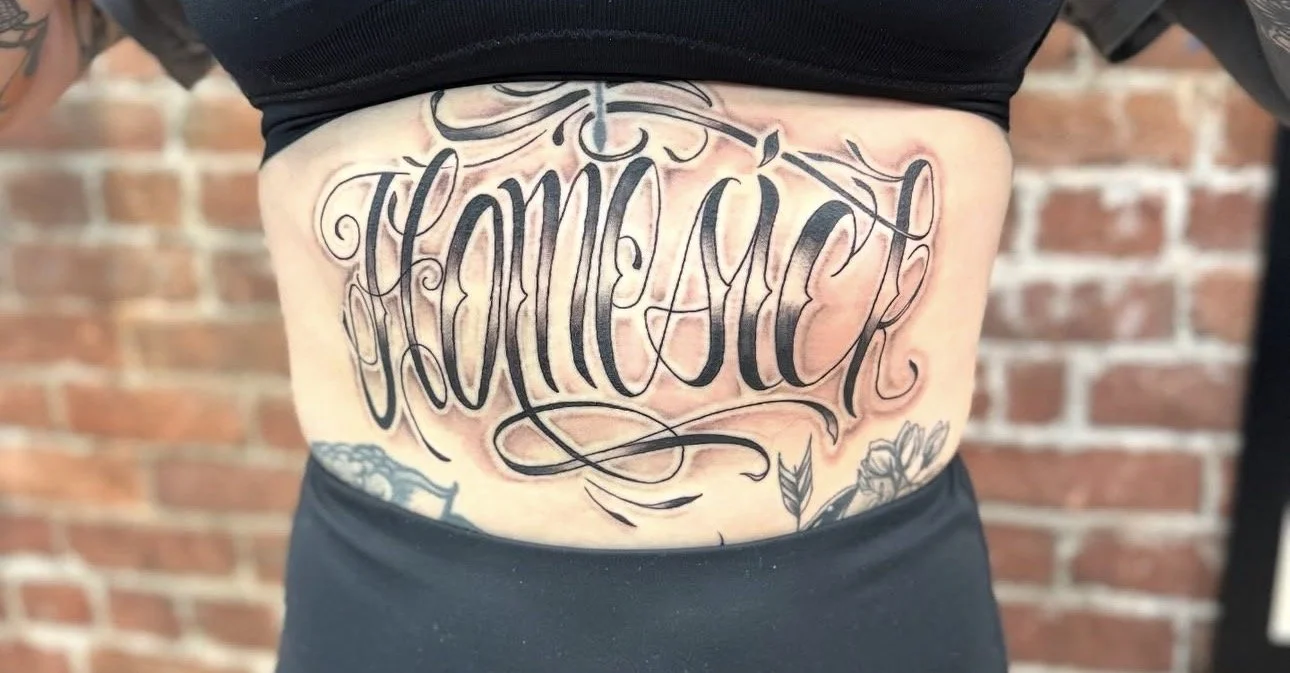 Tattoo reading 'Family' in ornate black script on person's stomach with floral and decorative elements