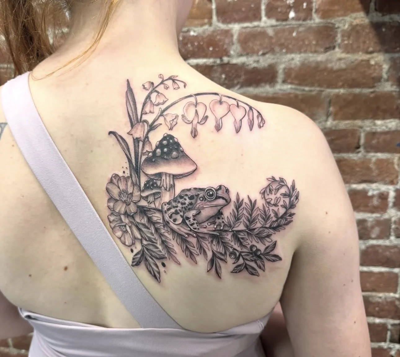 Tattoo of mushrooms, a frog, and flowers on a person's shoulder and chest with a brick wall background.