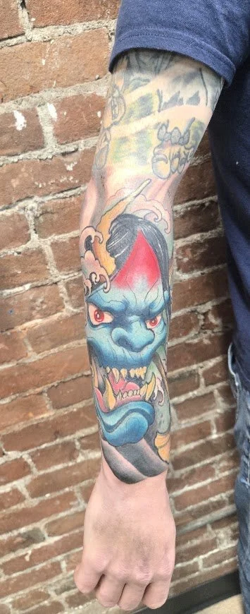 Tattoo of a blue demon face with red eyes and sharp teeth on a person's arm against a brick wall background.