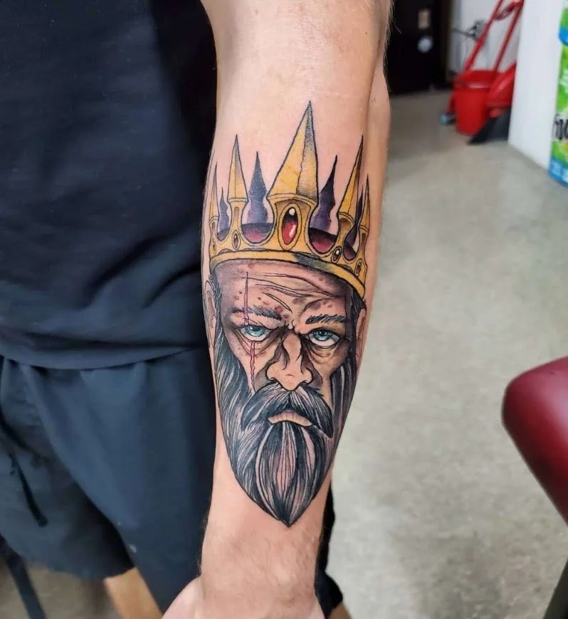 Tattoo of an elderly man with a long beard wearing a crown on his head.