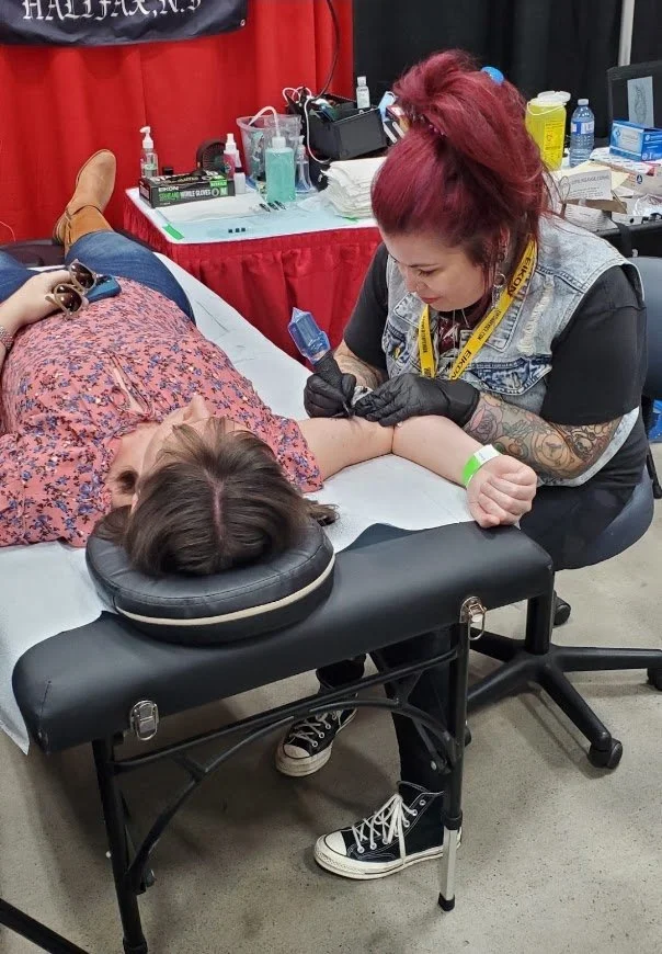 A woman with red hair, tattoos, and a yellow lanyard tattoo is tattooing a person's arm during a tattoo convention or event. The person getting tattooed is lying on a black massage table, wearing a pink floral dress, and is being treated on her left arm.