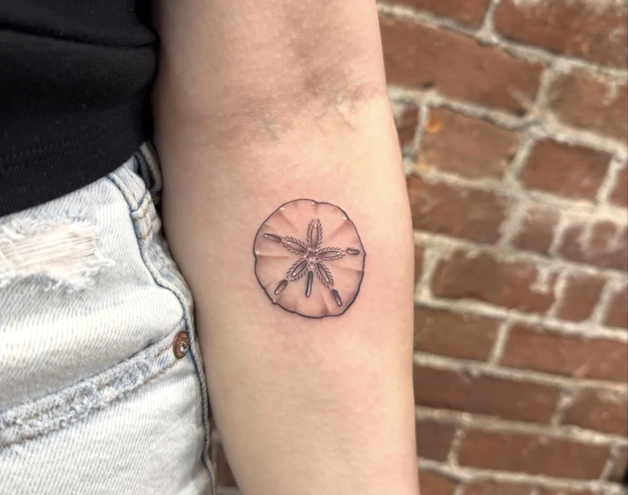 A tattoo of a lemon slice with a cannabis leaf in the center, located on a person's upper arm near the shoulder, with a brick wall in the background.