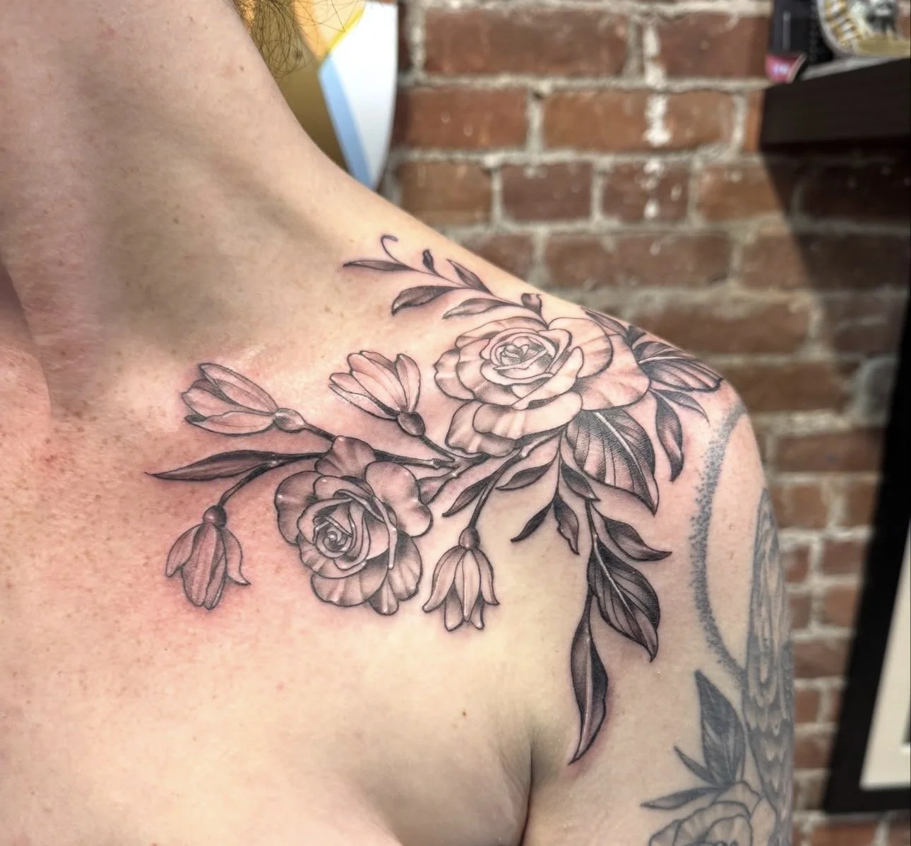 A black and gray floral tattoo on a person's shoulder and upper chest, featuring roses, leaves, and buds.