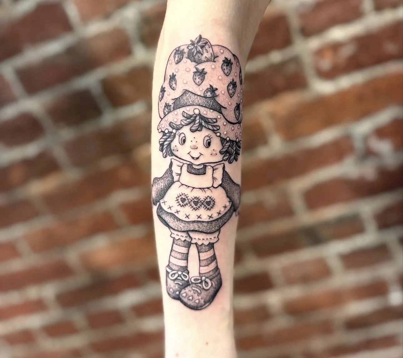 Black and white tattoo of a girl with curly hair wearing a strawberry hat, striped socks, and a dress with patchwork on the skirt, holding a teacup, in front of a brick wall.