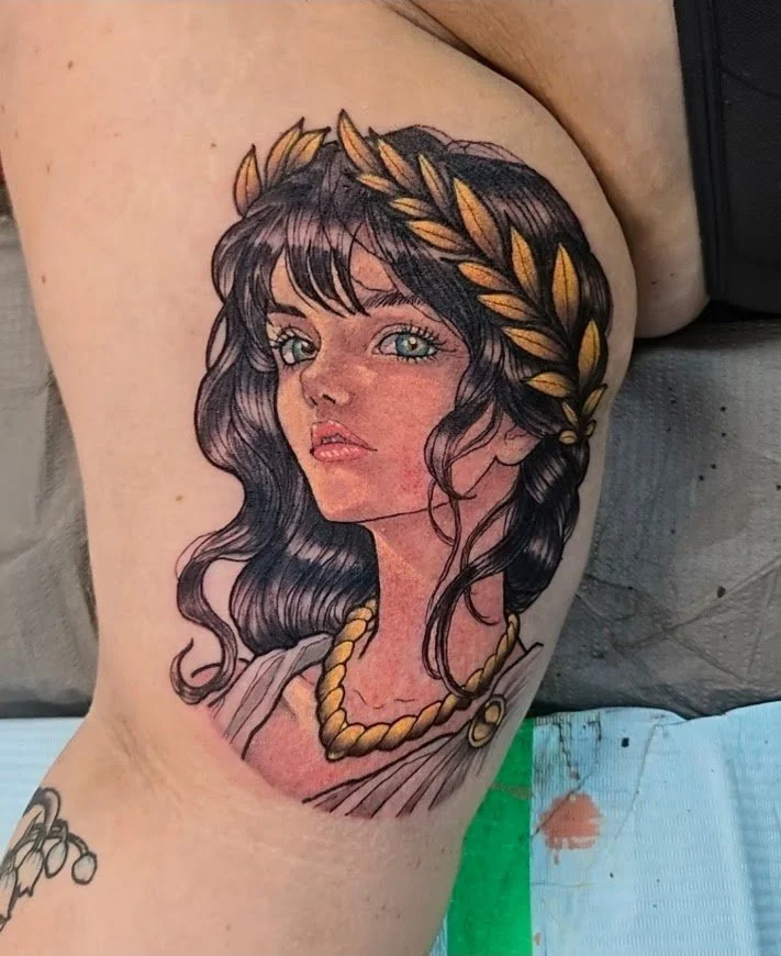 Colorful tattoo of a woman with dark, wavy hair and blue eyes, wearing a leaf crown and a gold necklace. aphrodite