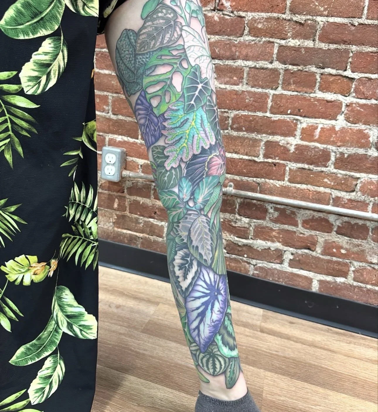 Close-up of a person's arm with a colorful botanical tattoo featuring various leaves and plants, standing against a brick wall with a wooden floor.