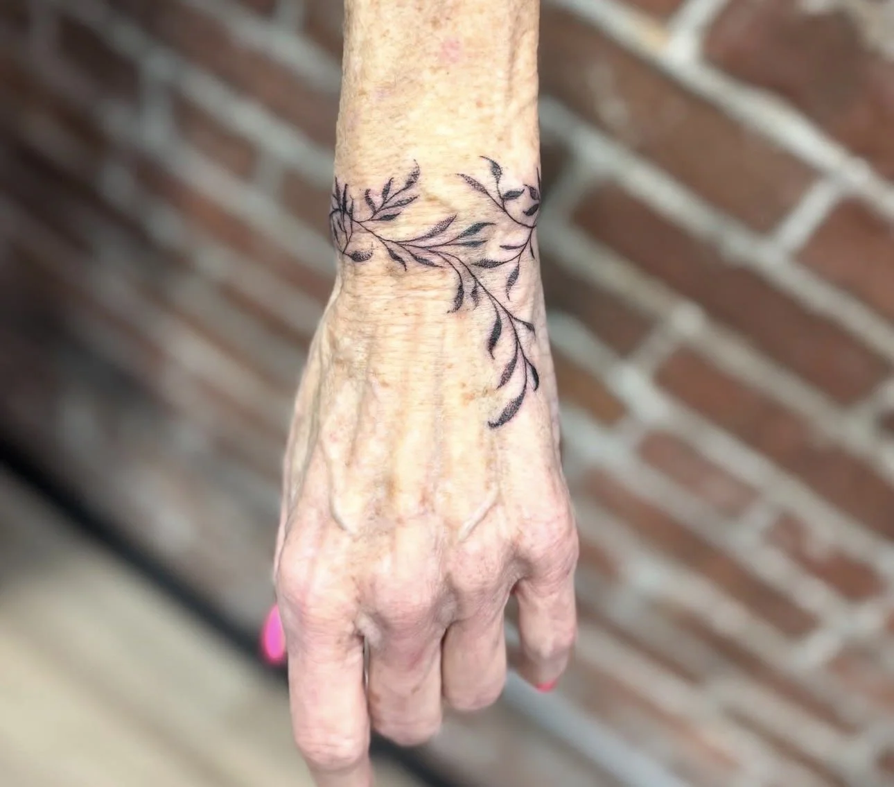 A person's hand with a black outline tattoo of leaves and branches on the wrist, against a brick wall background.