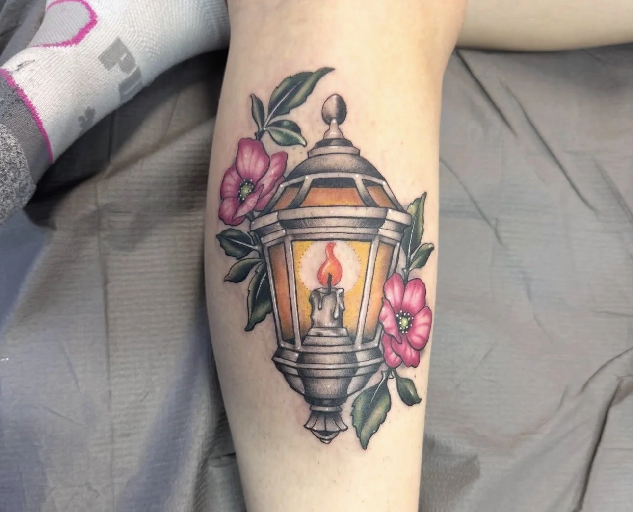 Tattoo of a street lantern with a candle inside, surrounded by pink flowers and green leaves.