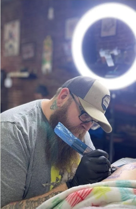 A tattoo artist with a beard, glasses, a baseball cap, and black gloves is tattooing someone at a tattoo studio. There is a lit ring light behind him and some artwork on the brick wall in the background. Greg Marsden